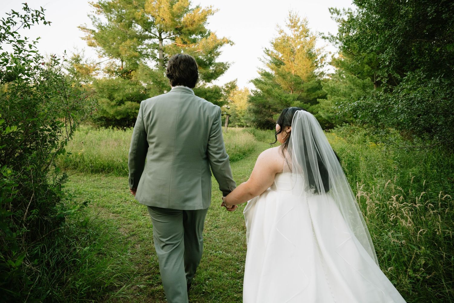 A newlywed couple walks hand in hand through a lush green meadow surrounded by autumn-touched trees.