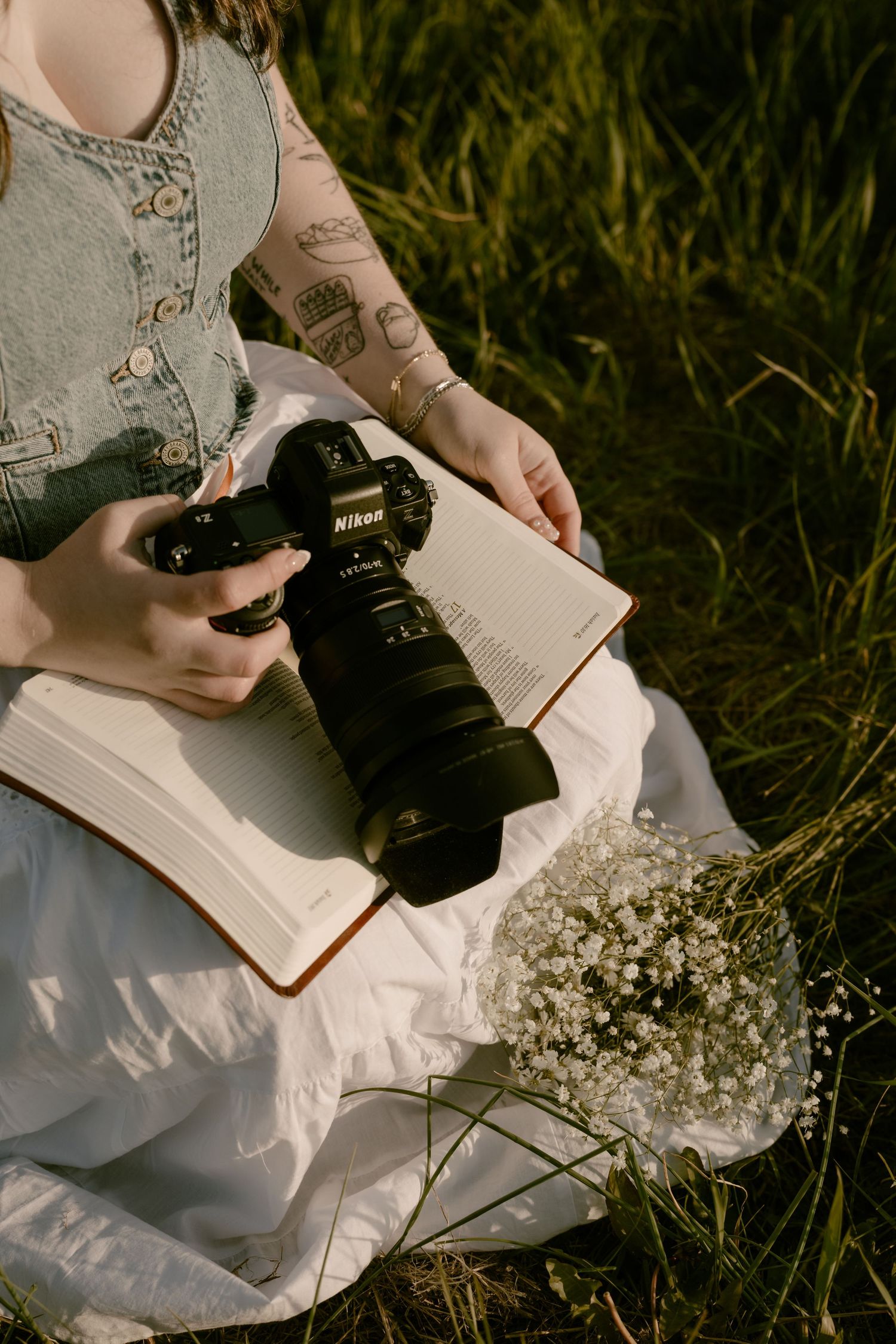 Close-up of woman holding Nikon camera and open book with baby's breath flowers in grass.