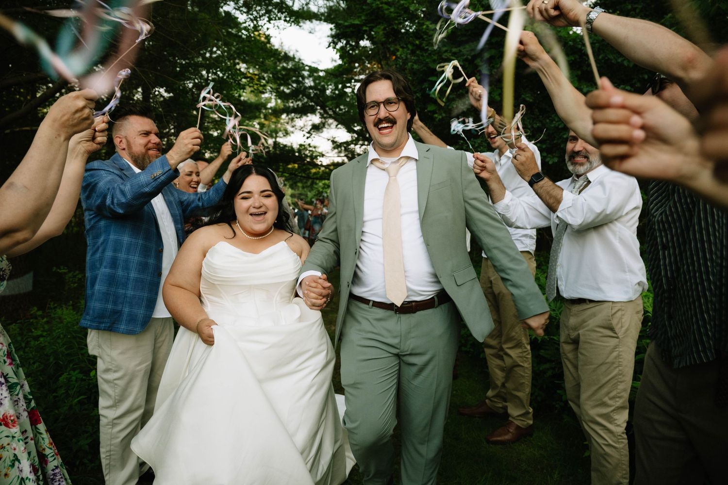 A joyful newlywed couple walks through a celebratory ribbon wand send-off surrounded by cheering guests.