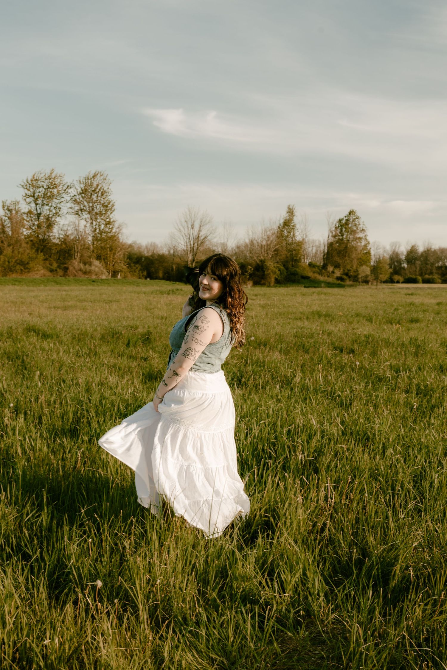 A woman in a flowing white skirt and denim jacket twirls in a sunlit green meadow.