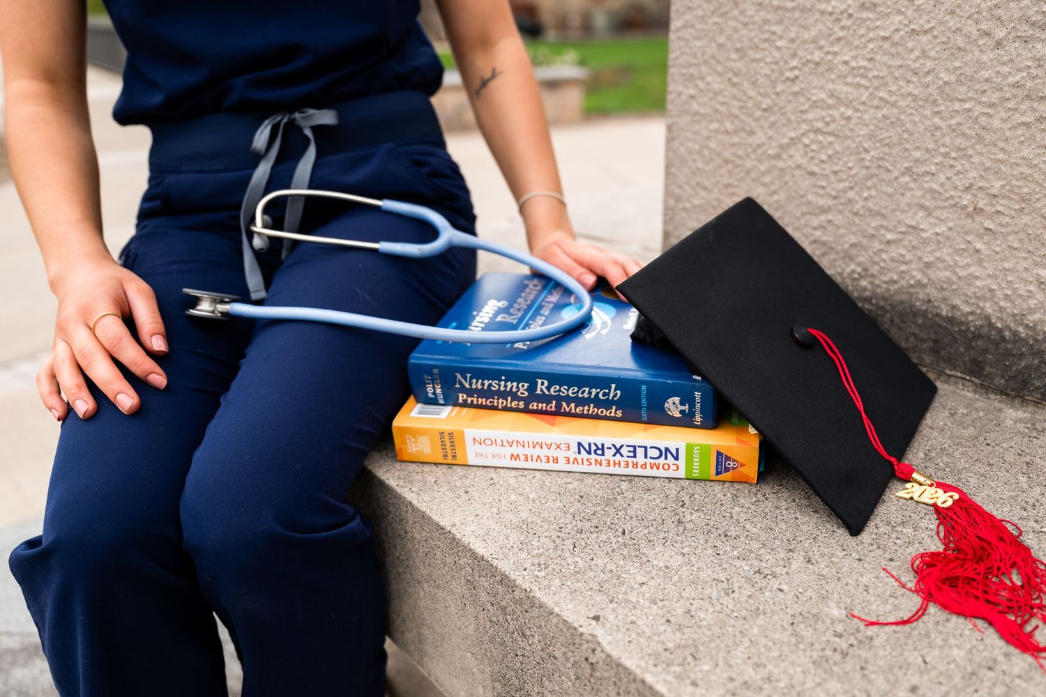 Nursing student in scrubs with stethoscope, textbooks, and graduation cap on stone steps.