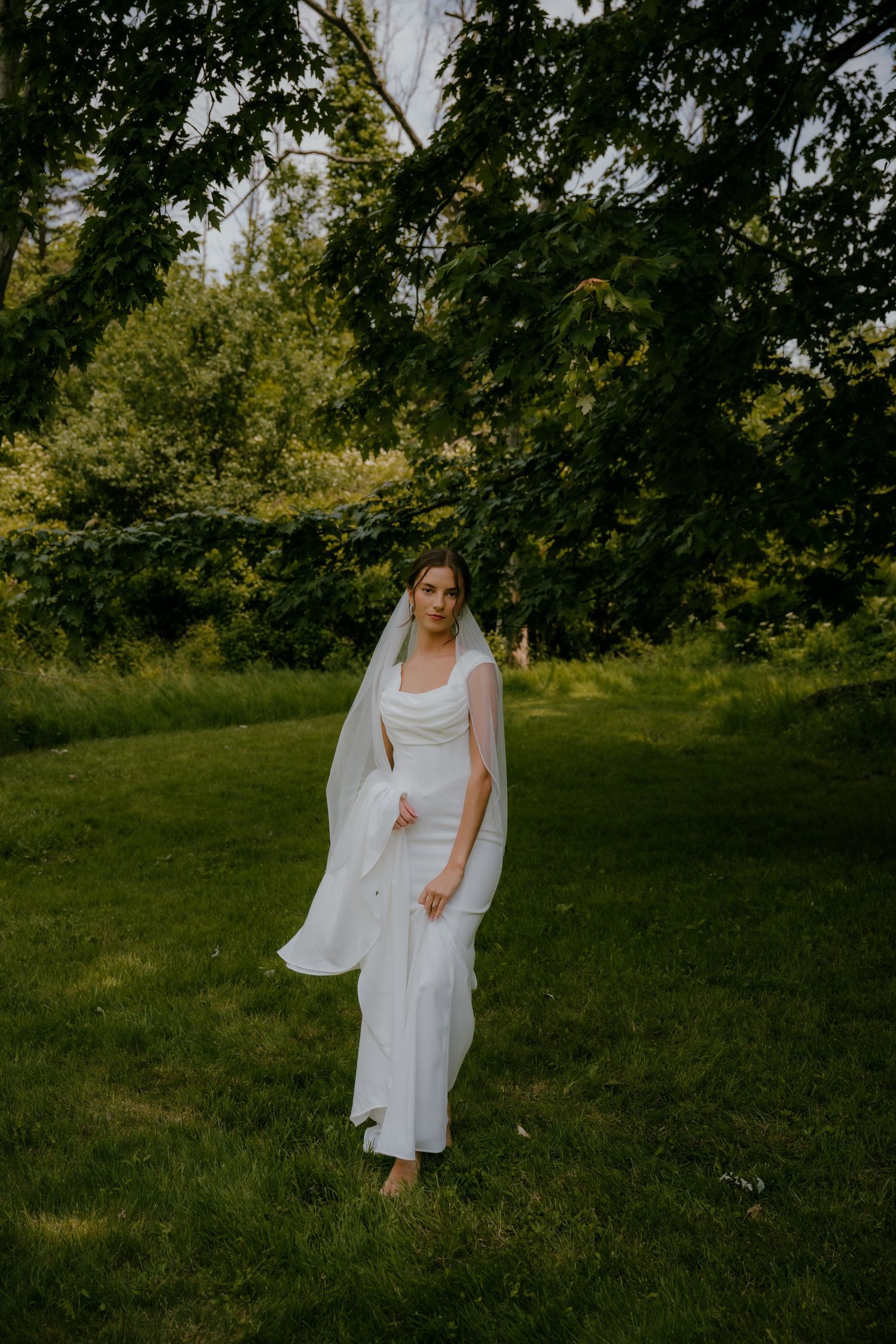 Bride in minimalist white dress and cathedral veil posing barefoot on green lawn under tree canopy.