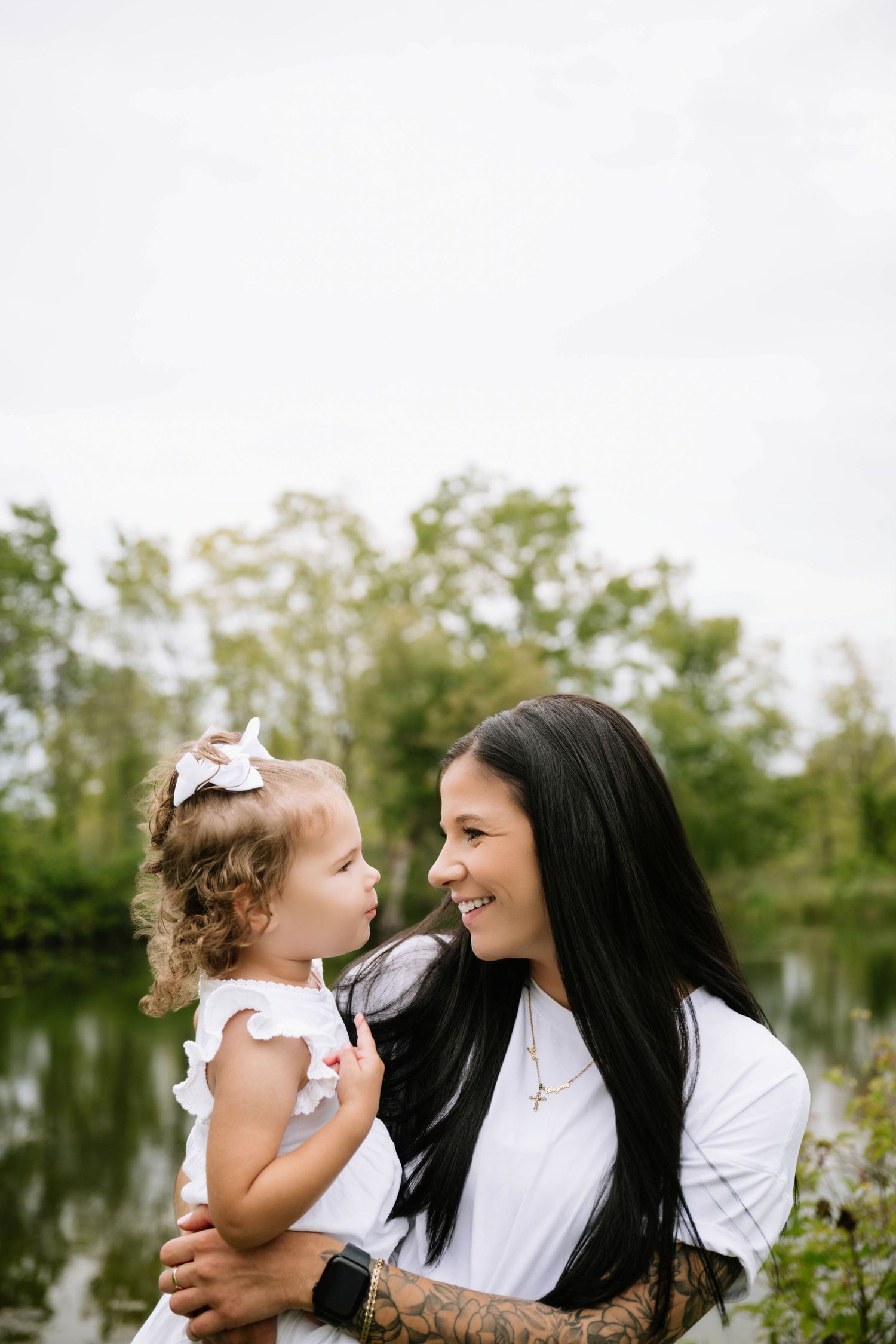 Smiling dark-haired mother holds her curly-haired toddler daughter near a peaceful pond.