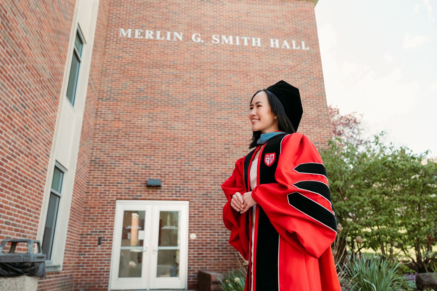 Woman in red doctoral graduation gown standing outside Merlin G. Smith Hall brick building.