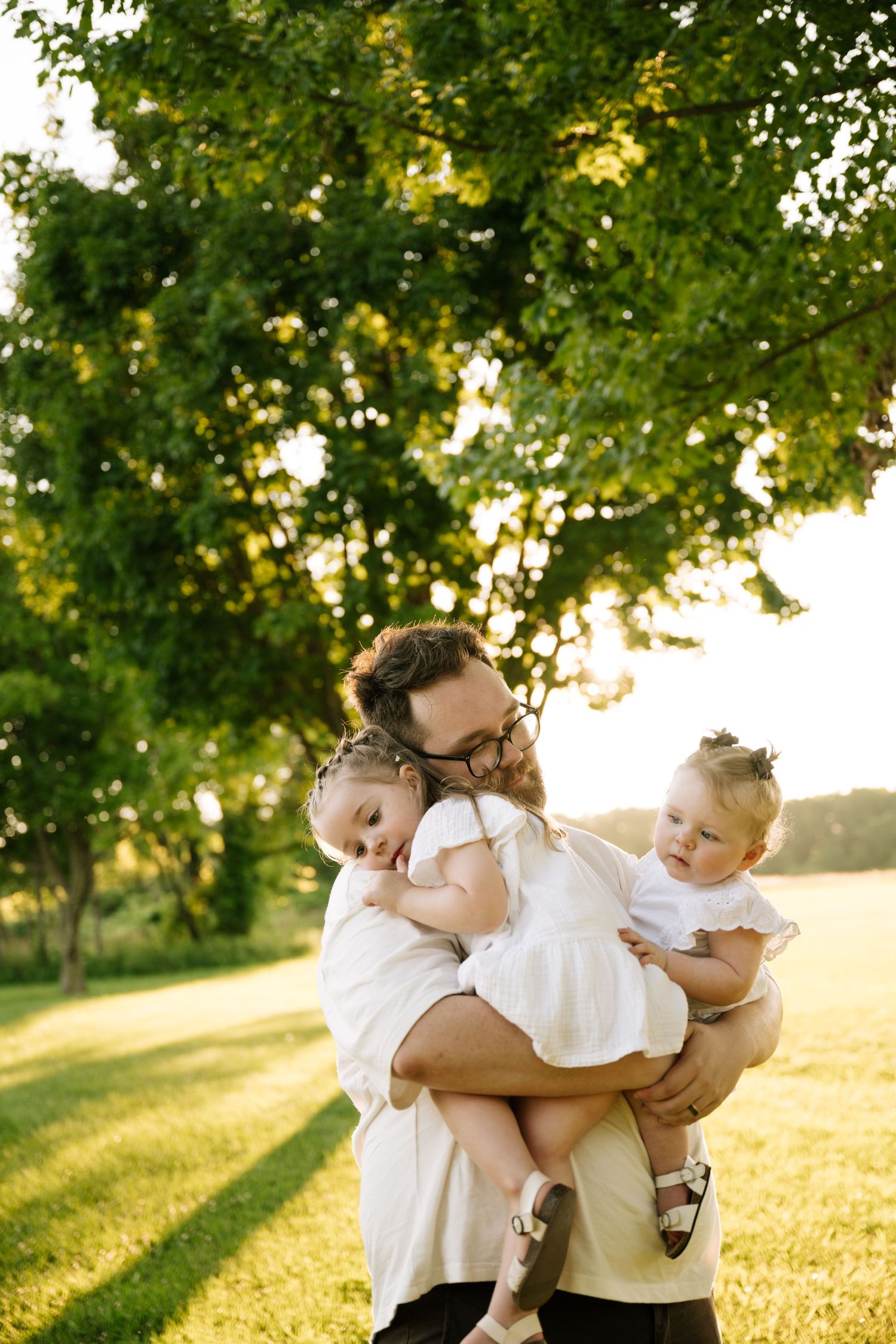 Father with glasses holding two young daughters in white dresses in a sunny green park.