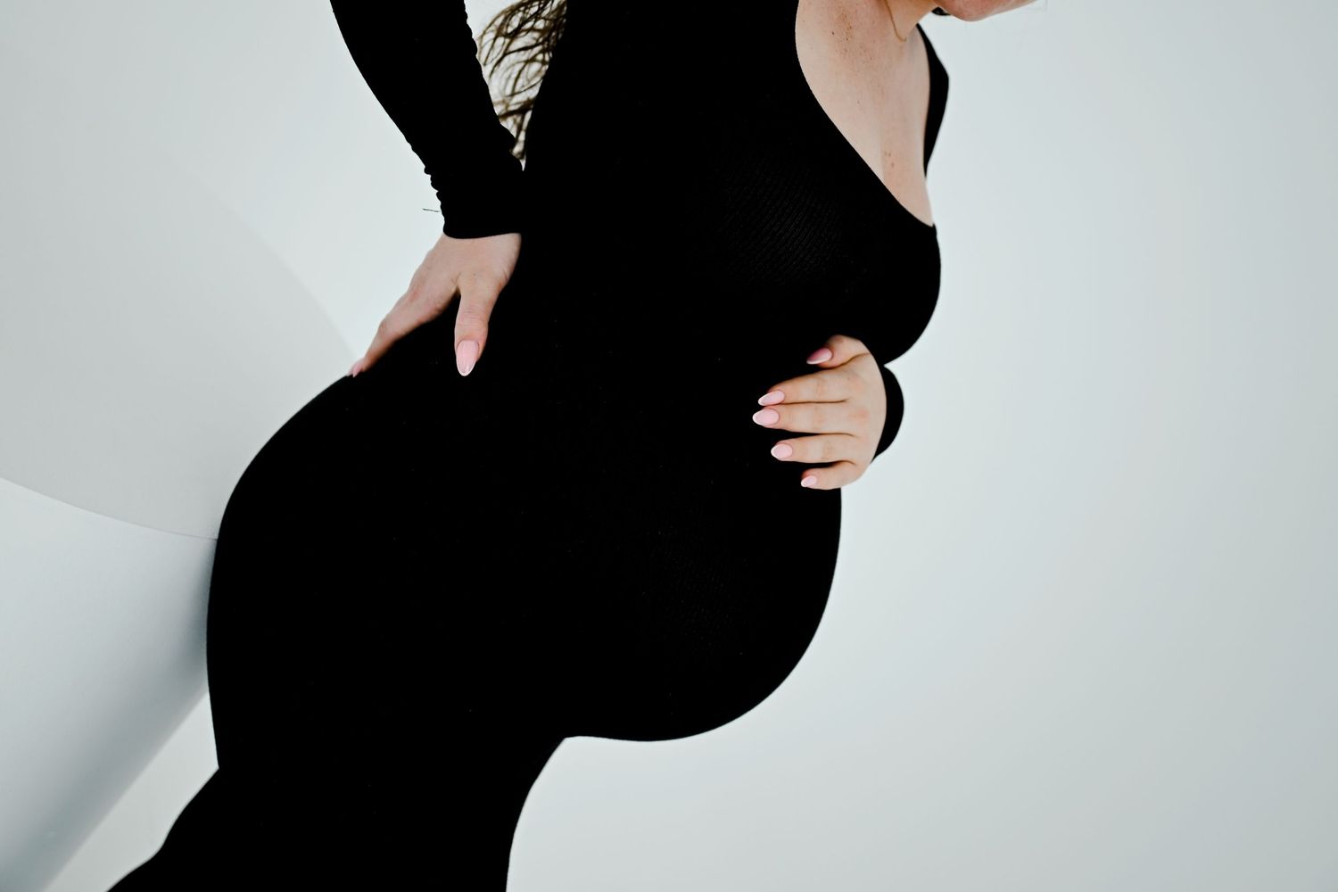 Side profile of pregnant woman in black dress highlighting baby bump against light background.