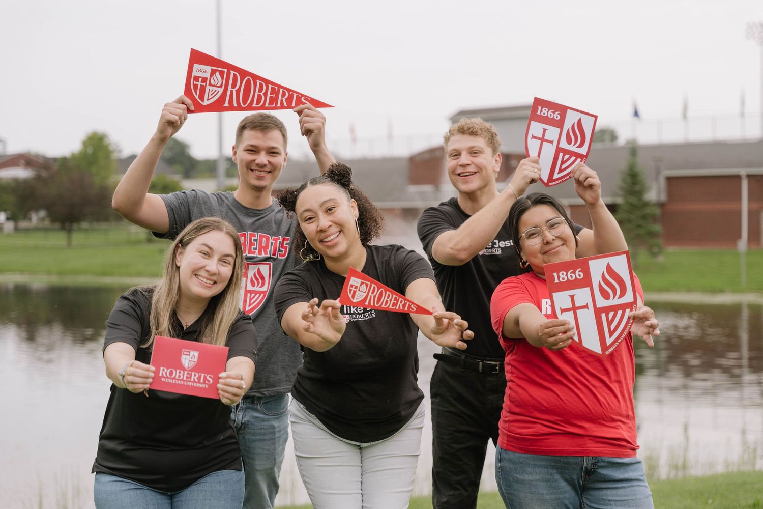Five college students smile and hold red Roberts Wesleyan University pennants and flags outdoors.