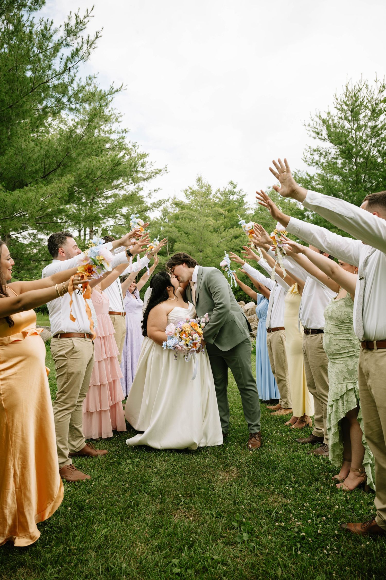 Bride and groom share kiss during outdoor wedding exit tunnel with guests raising hands in celebration.