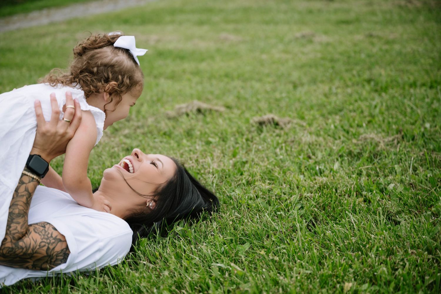 Mother lying on grass laughing while lifting her toddler daughter in white dress overhead.