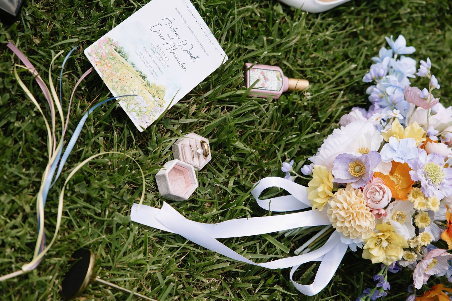 Wedding flat lay with invitation, ring boxes, and colorful bridal bouquet on green grass.