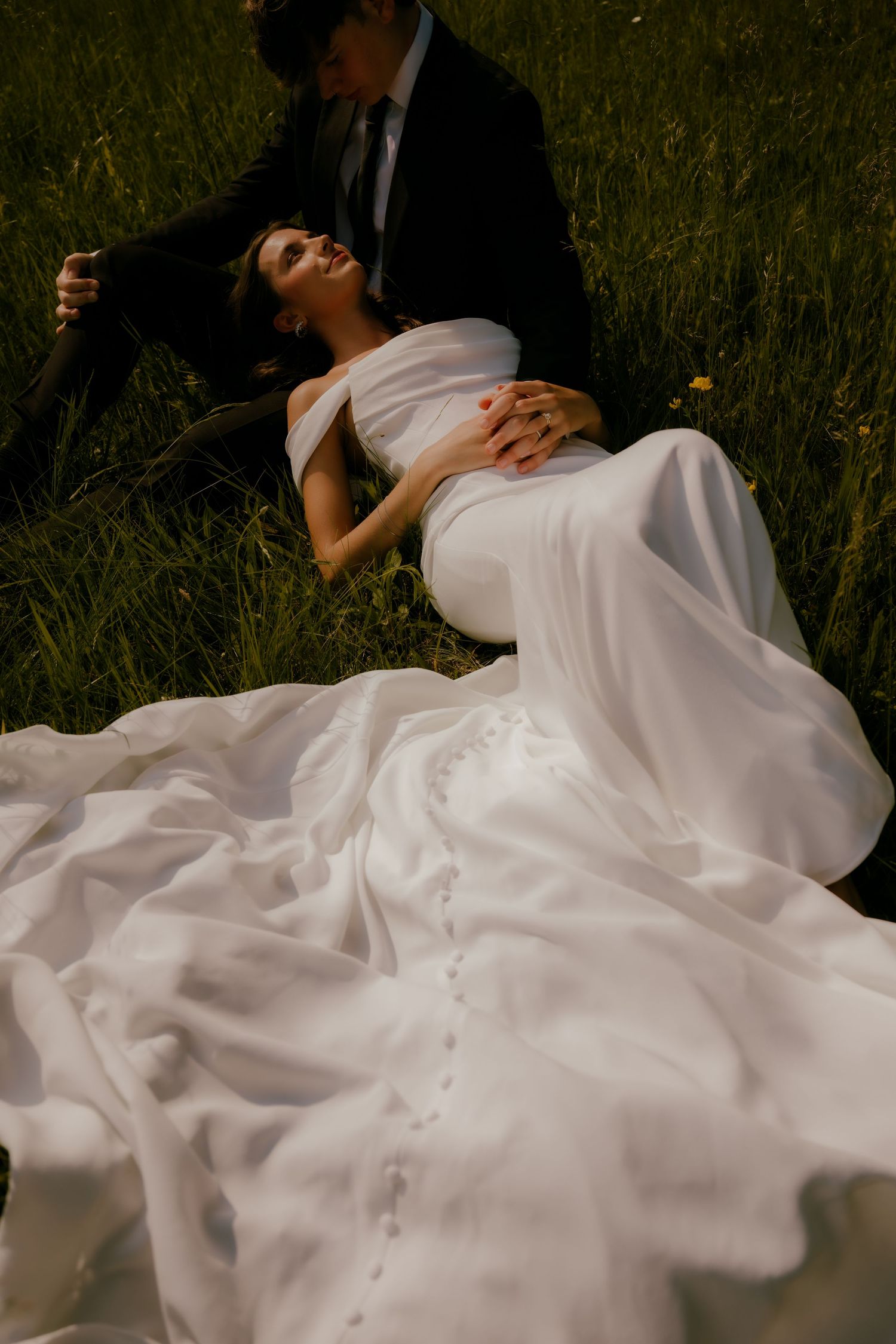 Bride in elegant white gown reclining on grass with groom in dark suit at outdoor wedding.