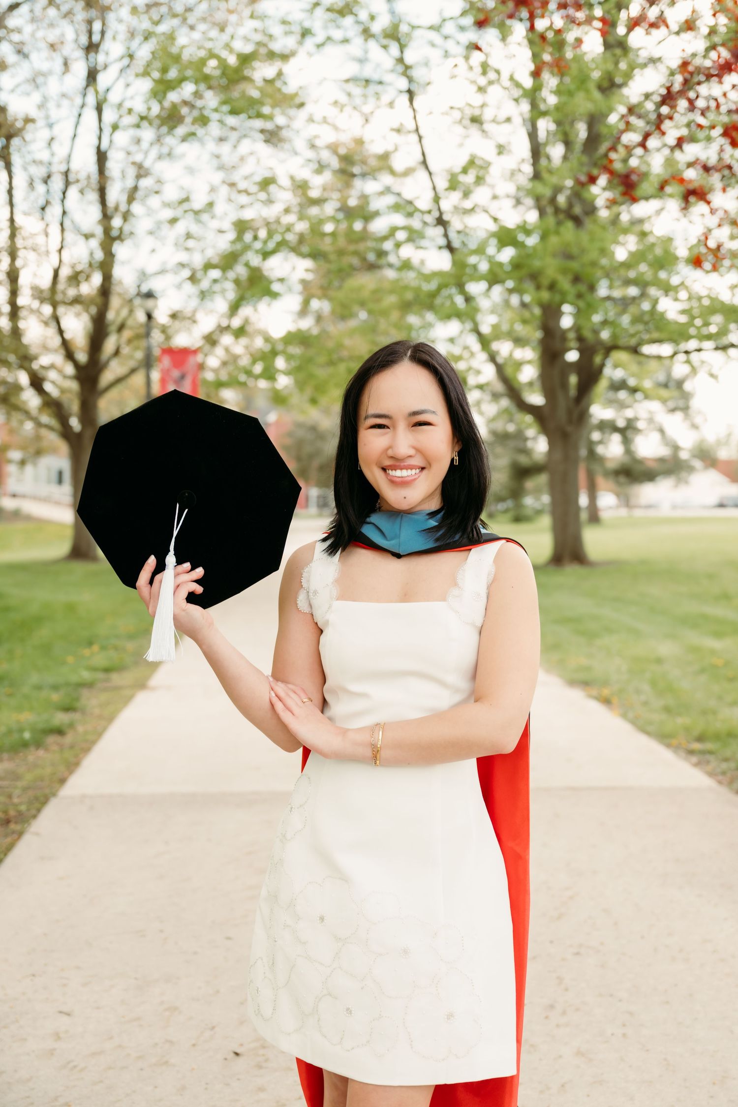 Smiling woman in white dress holding graduation cap outdoors on a tree-lined campus path.