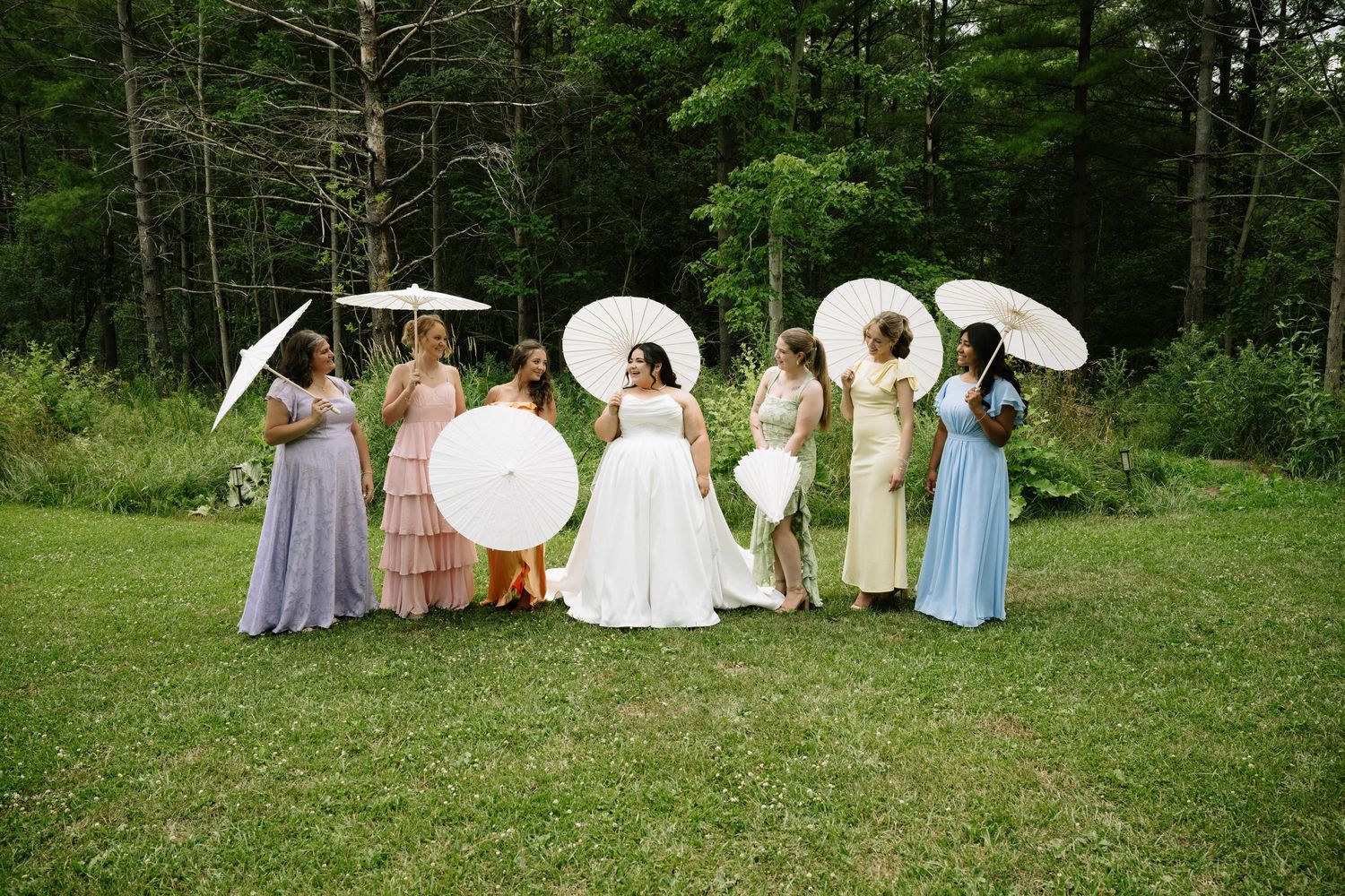 Bride and bridesmaids in colorful mismatched dresses holding white parasols in a garden.