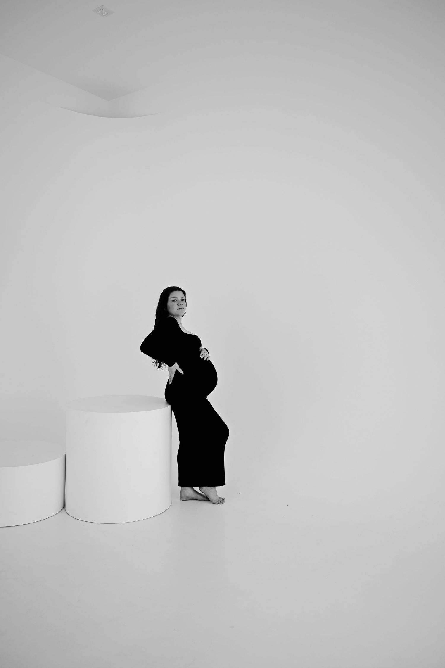 Black and white photo of pregnant woman in black dress posing in minimalist white studio.
