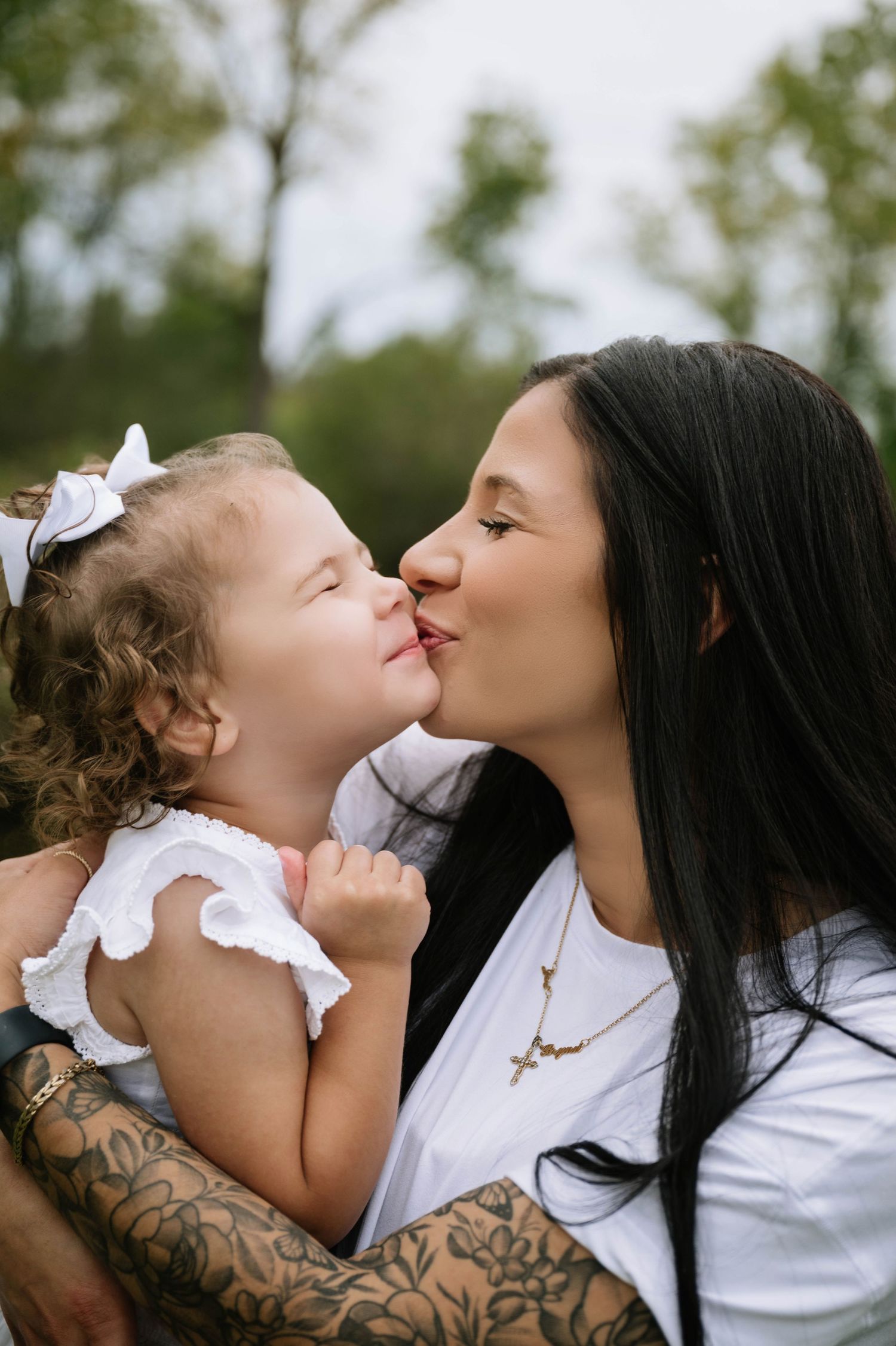 A tattooed mother shares a tender kiss with her curly-haired toddler daughter outdoors in nature.