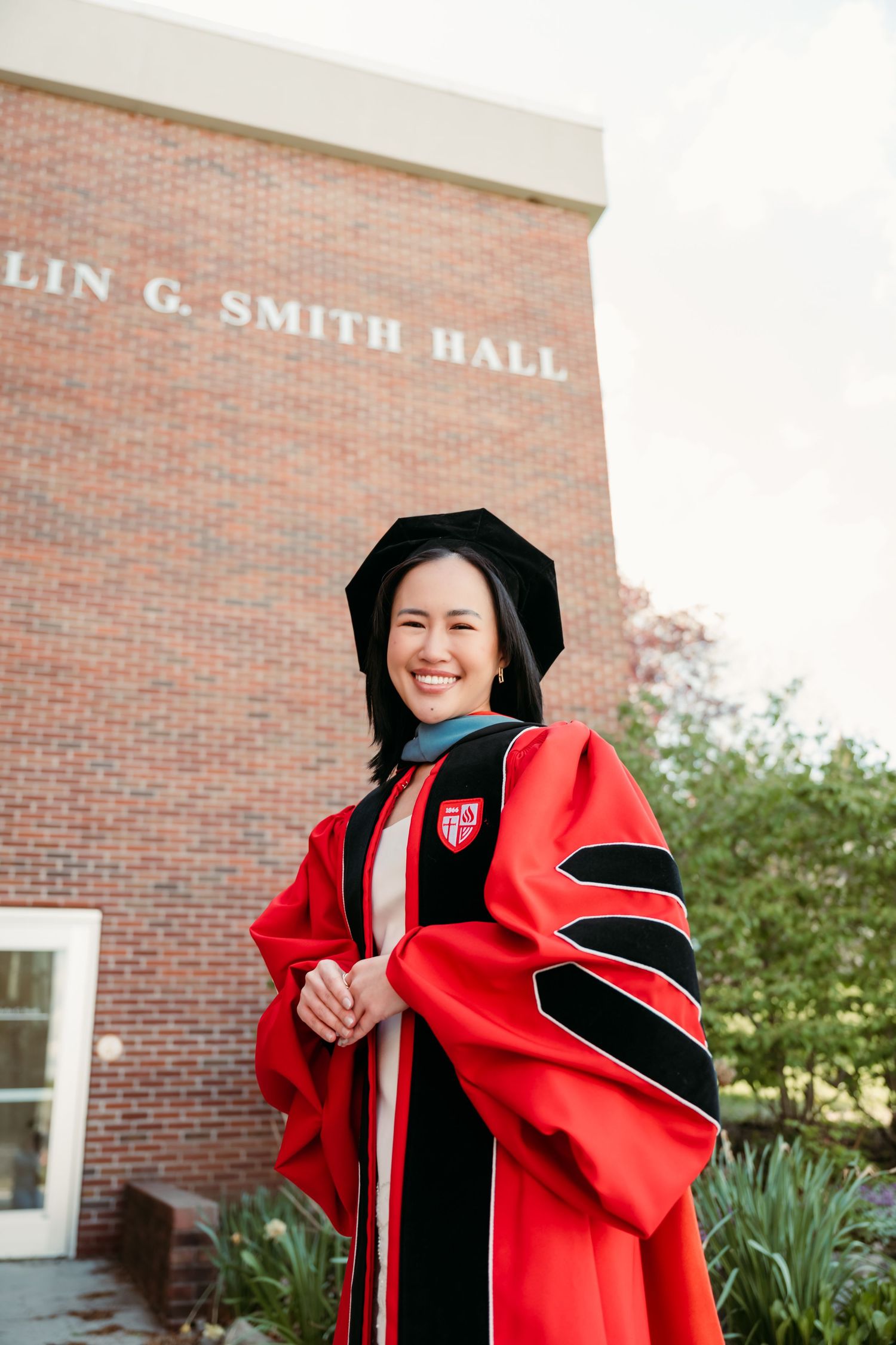 Proud graduate in red doctoral regalia smiles standing before Lin G. Smith Hall brick building.