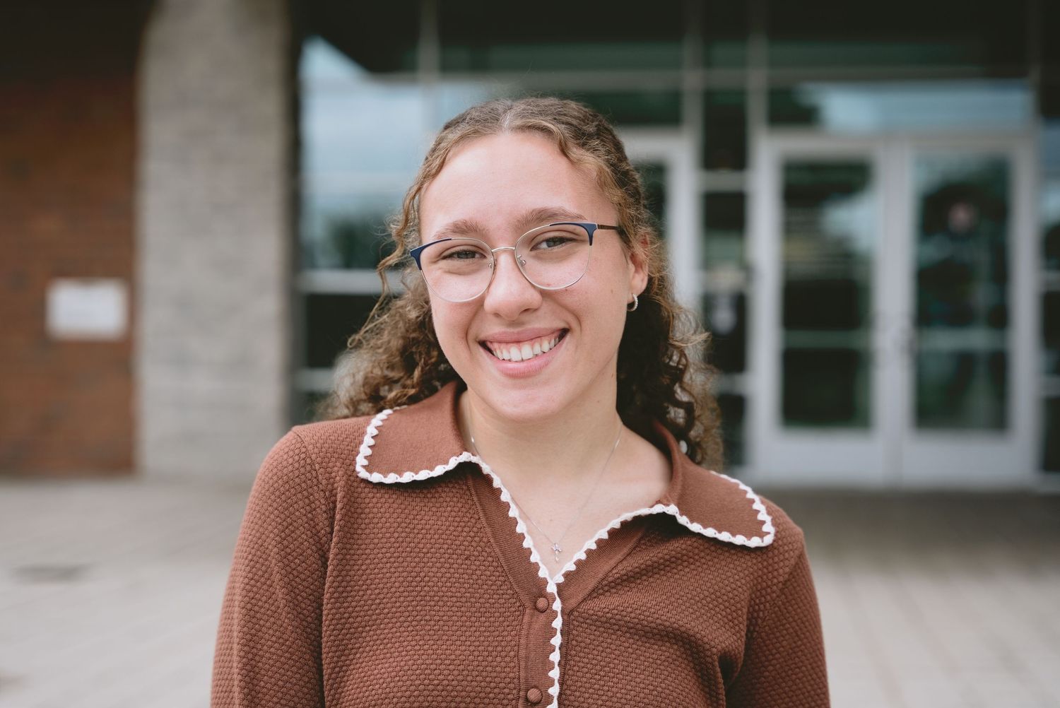 A smiling young woman with curly hair and glasses wears a brown cardigan outside a campus building.