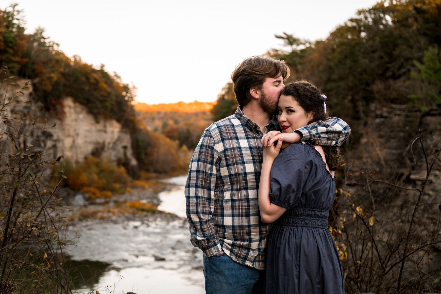 Couple embracing at sunset overlooking a scenic river gorge surrounded by autumn foliage.
