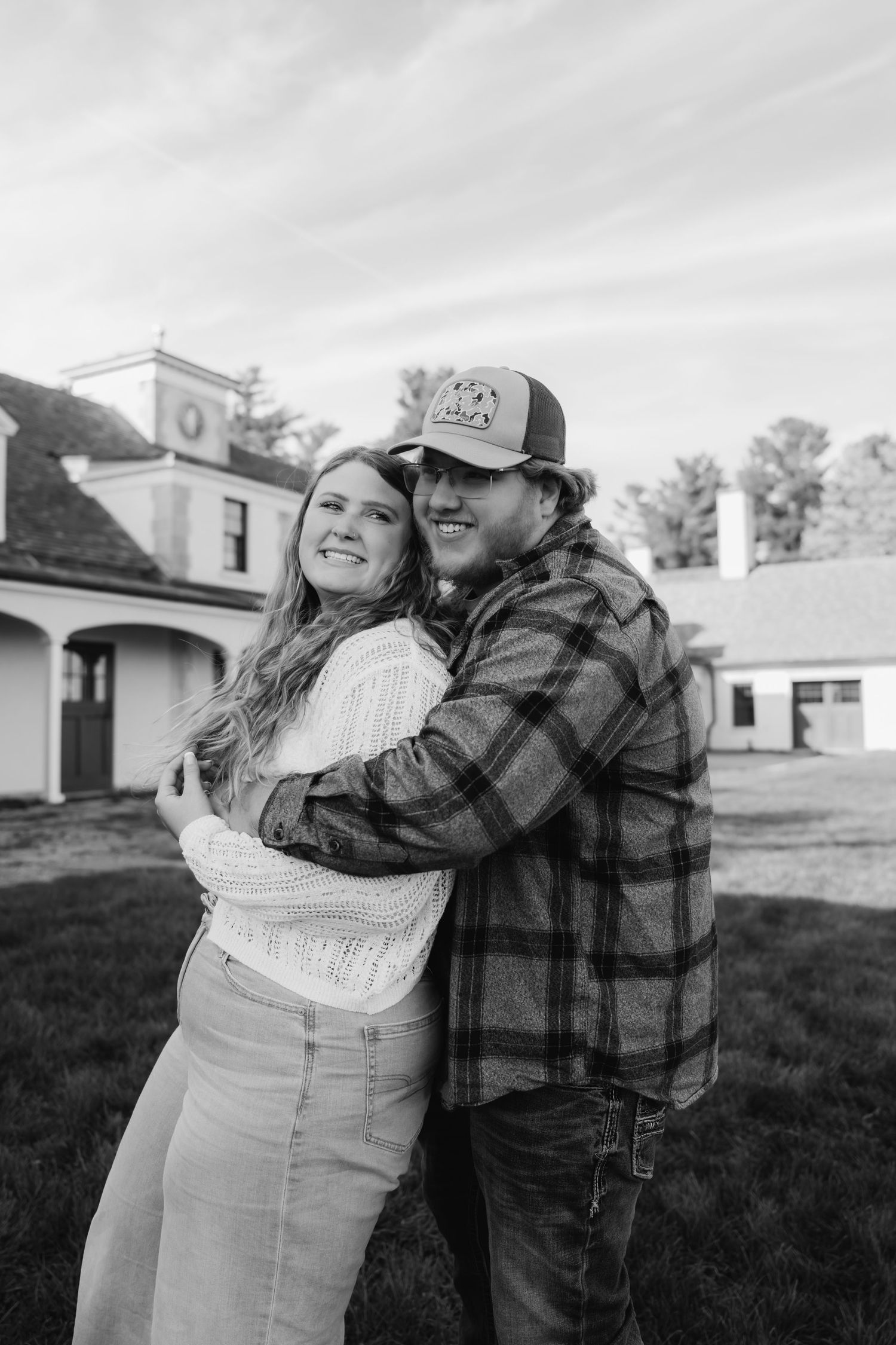 Black and white photo of smiling couple embracing outside a white building on a cloudy day.