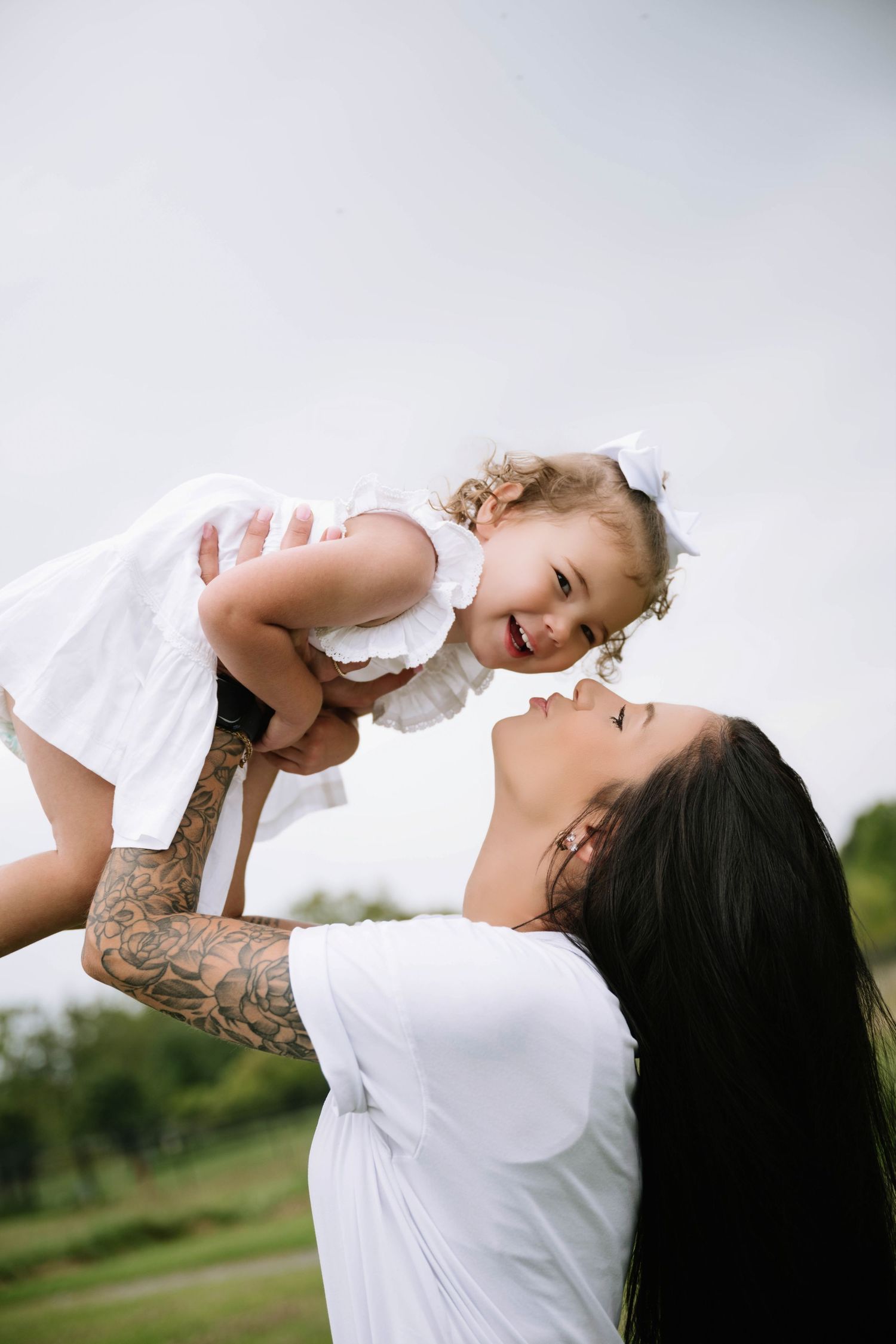 Tattooed mother lifting happy toddler daughter in white dress above her head outdoors.