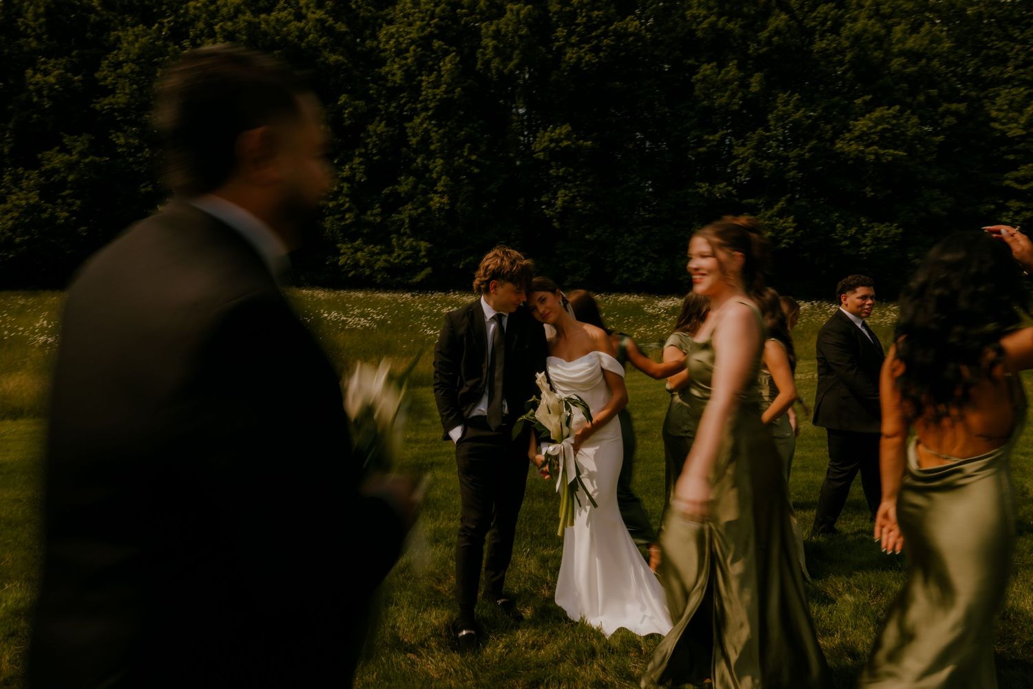 Bride and groom walking with wedding party across a sunlit green field at golden hour.