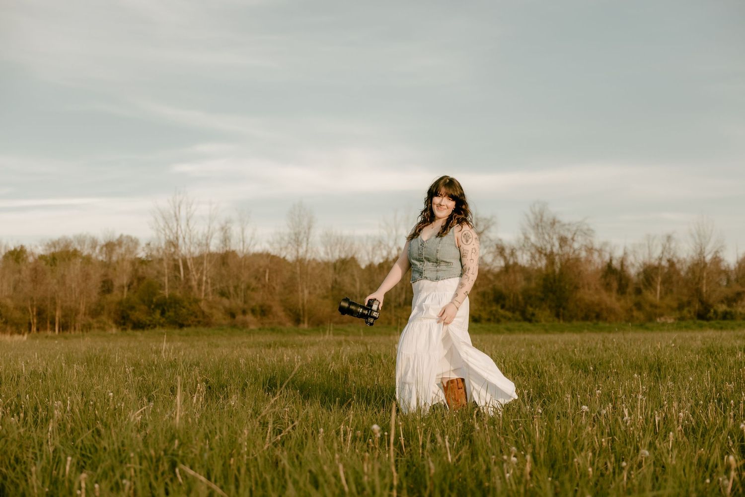 Woman in white skirt and green top walks through a sunlit green field holding a camera.