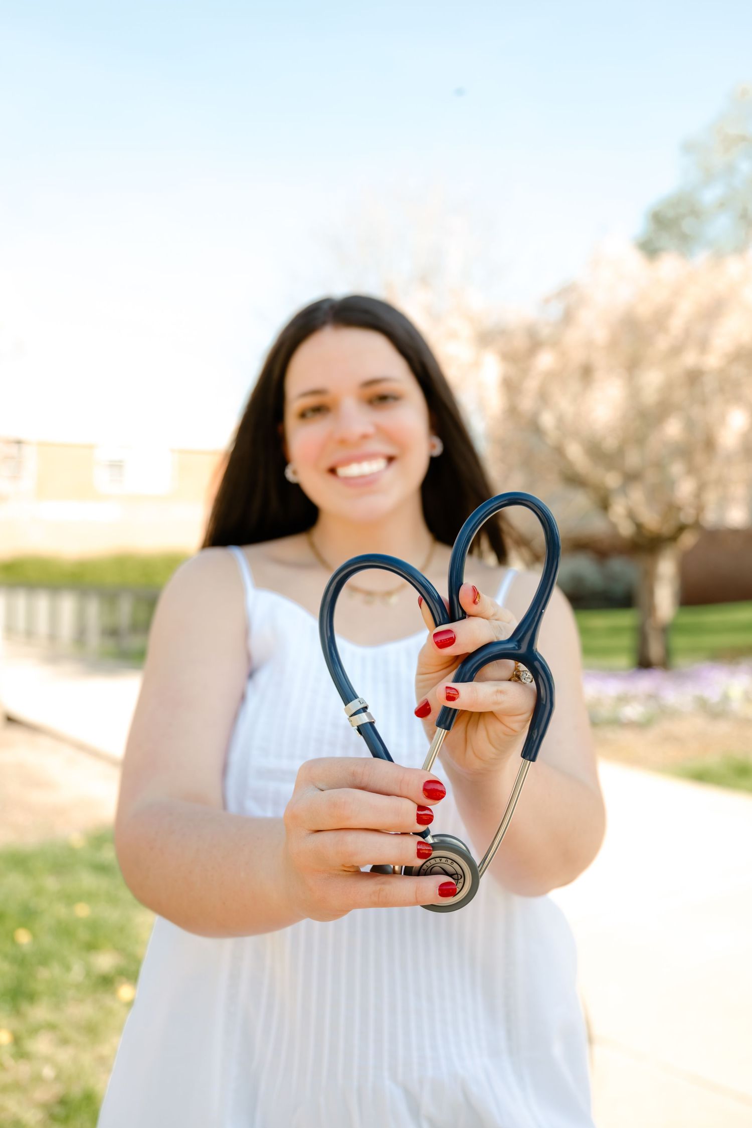 Woman in white dress smiling and holding a stethoscope shaped into a heart outdoors.