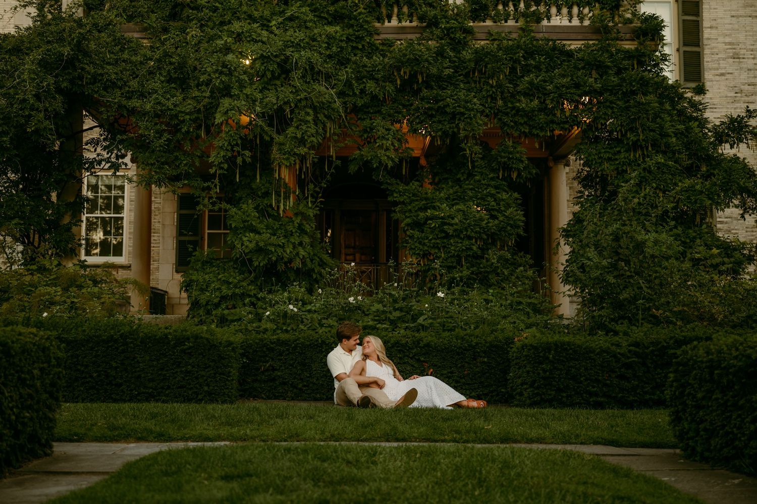 Couple in white attire sitting together on manicured lawn in front of an ivy-covered estate at dusk.