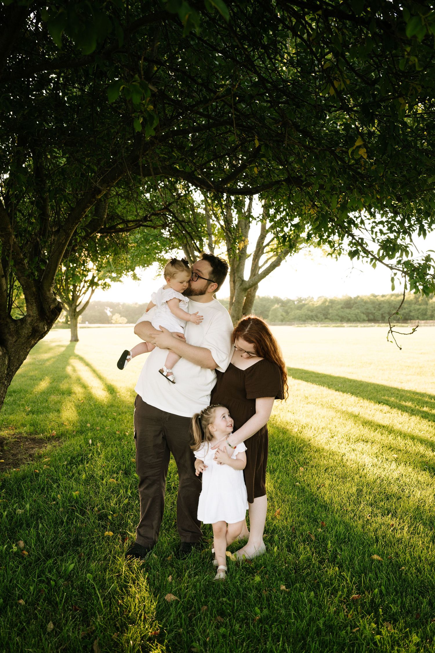 A bearded father kisses his baby while his pregnant wife and toddler stand beside him outdoors.