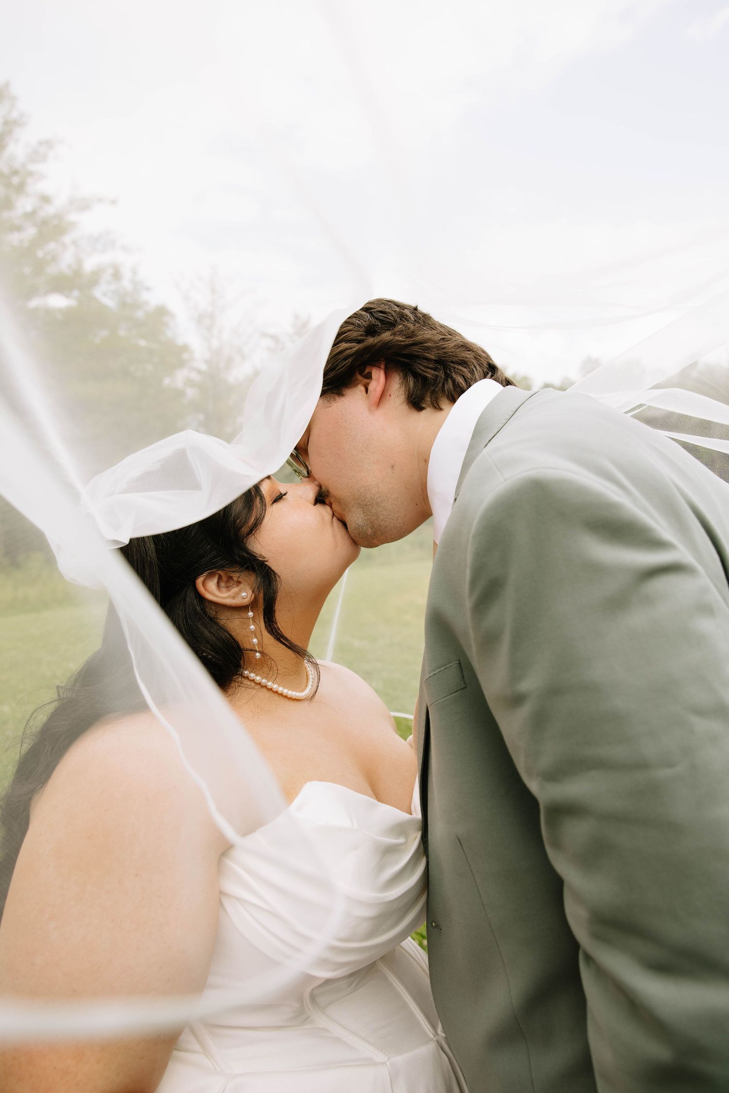 Bride and groom sharing a kiss under a flowing white veil outdoors on a cloudy day.