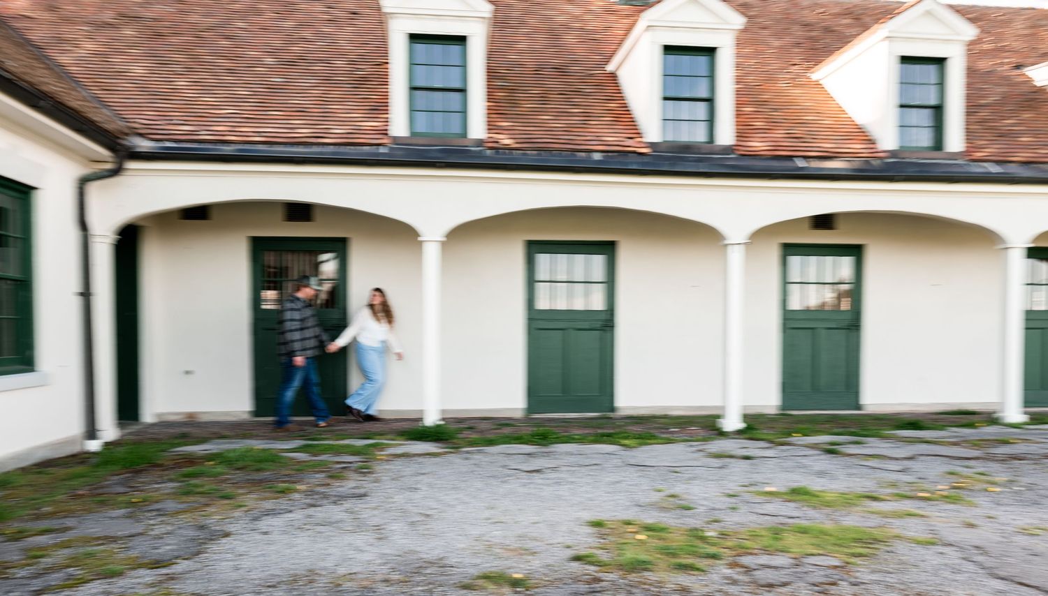 A couple walking together through a white arched building with green doors on a cobblestone courtyard.