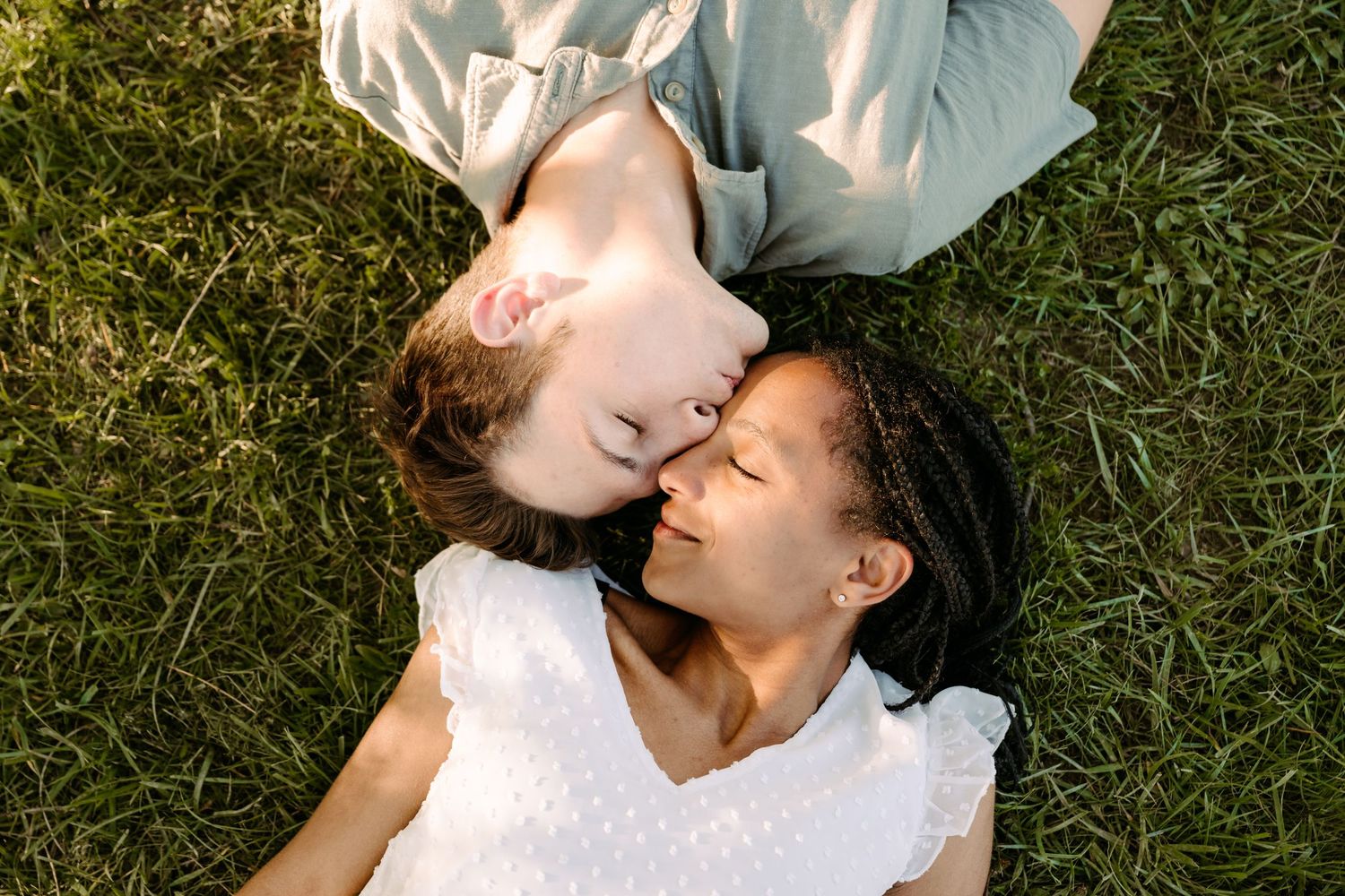 Two women lying on green grass face to face with eyes closed, sharing an intimate romantic moment outdoors.