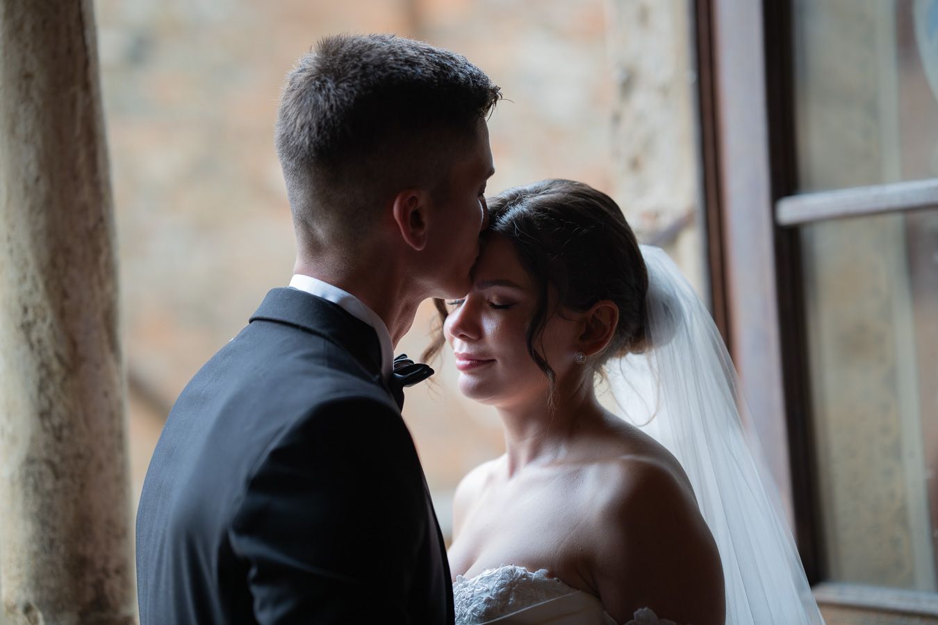 Couple standing closely together beside a window in an elegant indoor setting in Volterra
