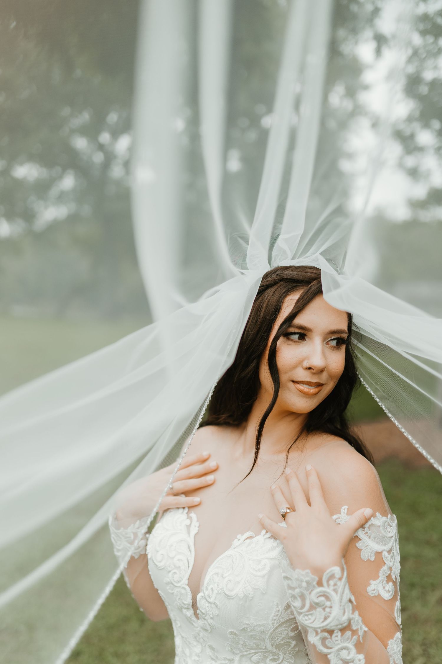 A bride in a delicate lace wedding gown poses with flowing white veil in a dreamy outdoor setting. Spartanburg Wedding photo