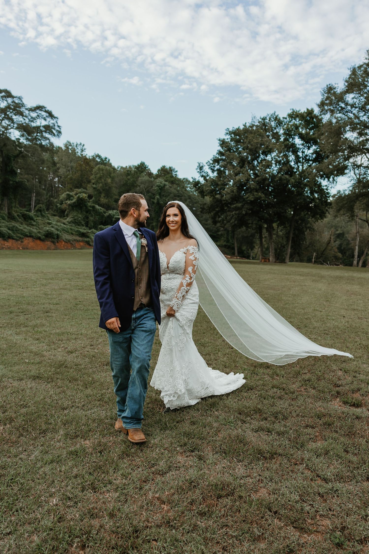 A couple in wedding attire stands together on a grassy field with trees and cloudy sky in the background.