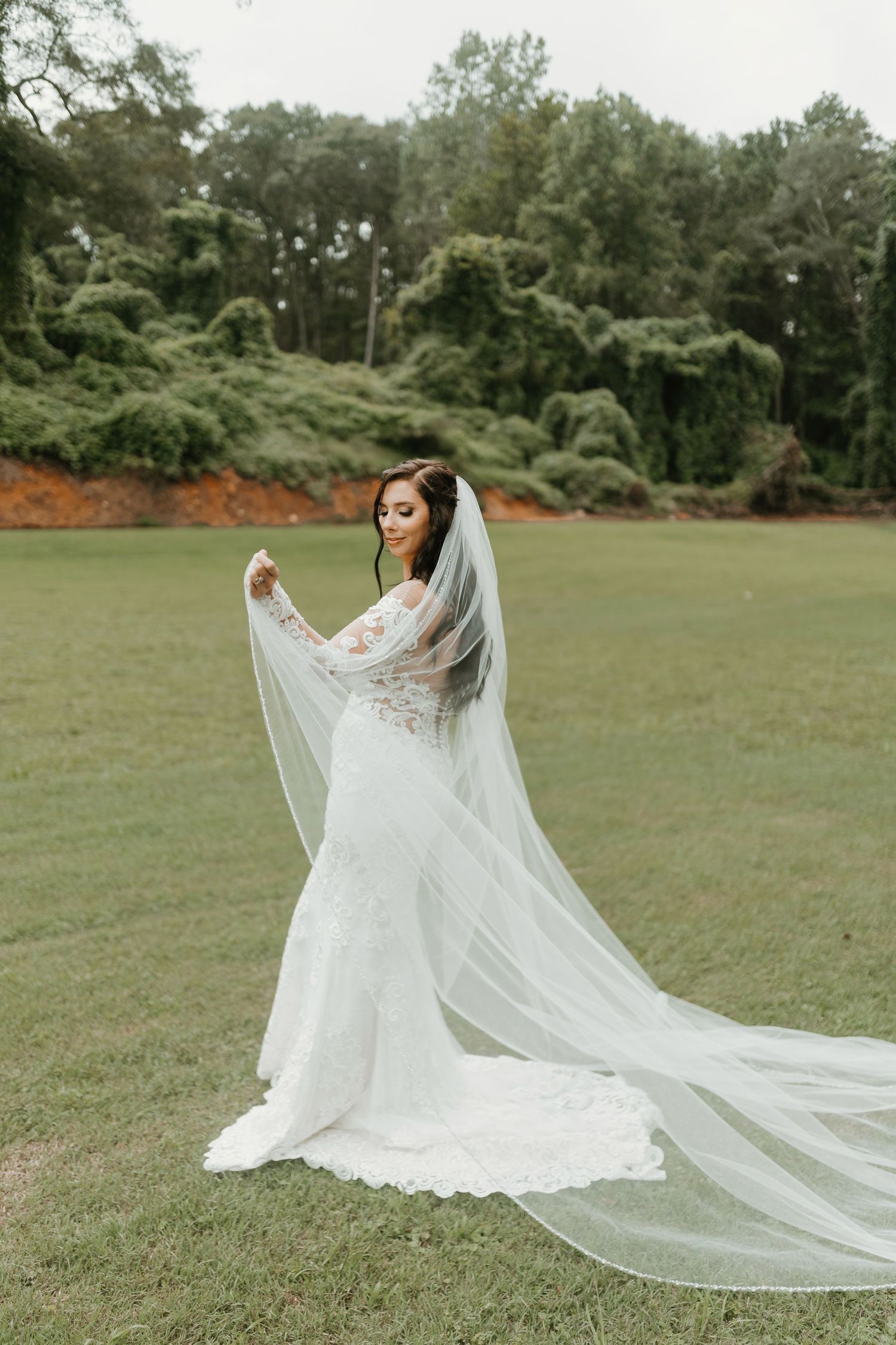 A bride in a flowing lace wedding gown with cathedral veil stands gracefully in a lush green field.