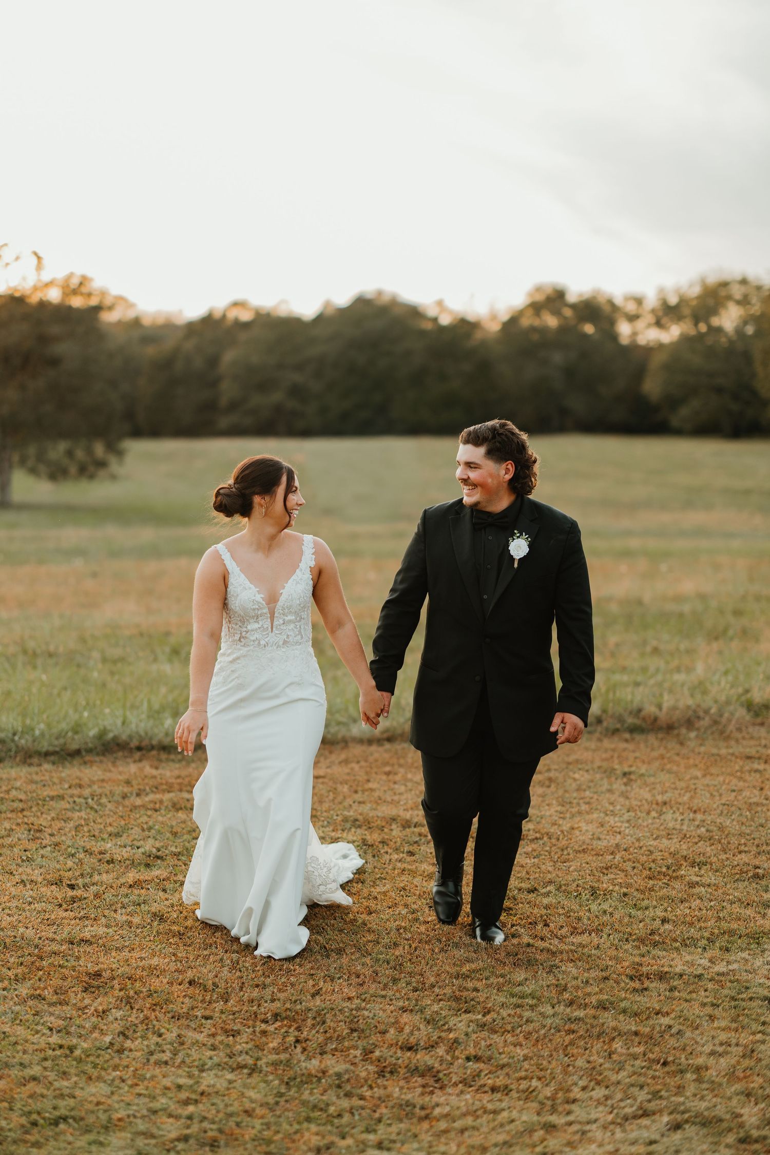 A couple in wedding attire walks together across a golden field at sunset wedding with Greenville Wedding Photographer