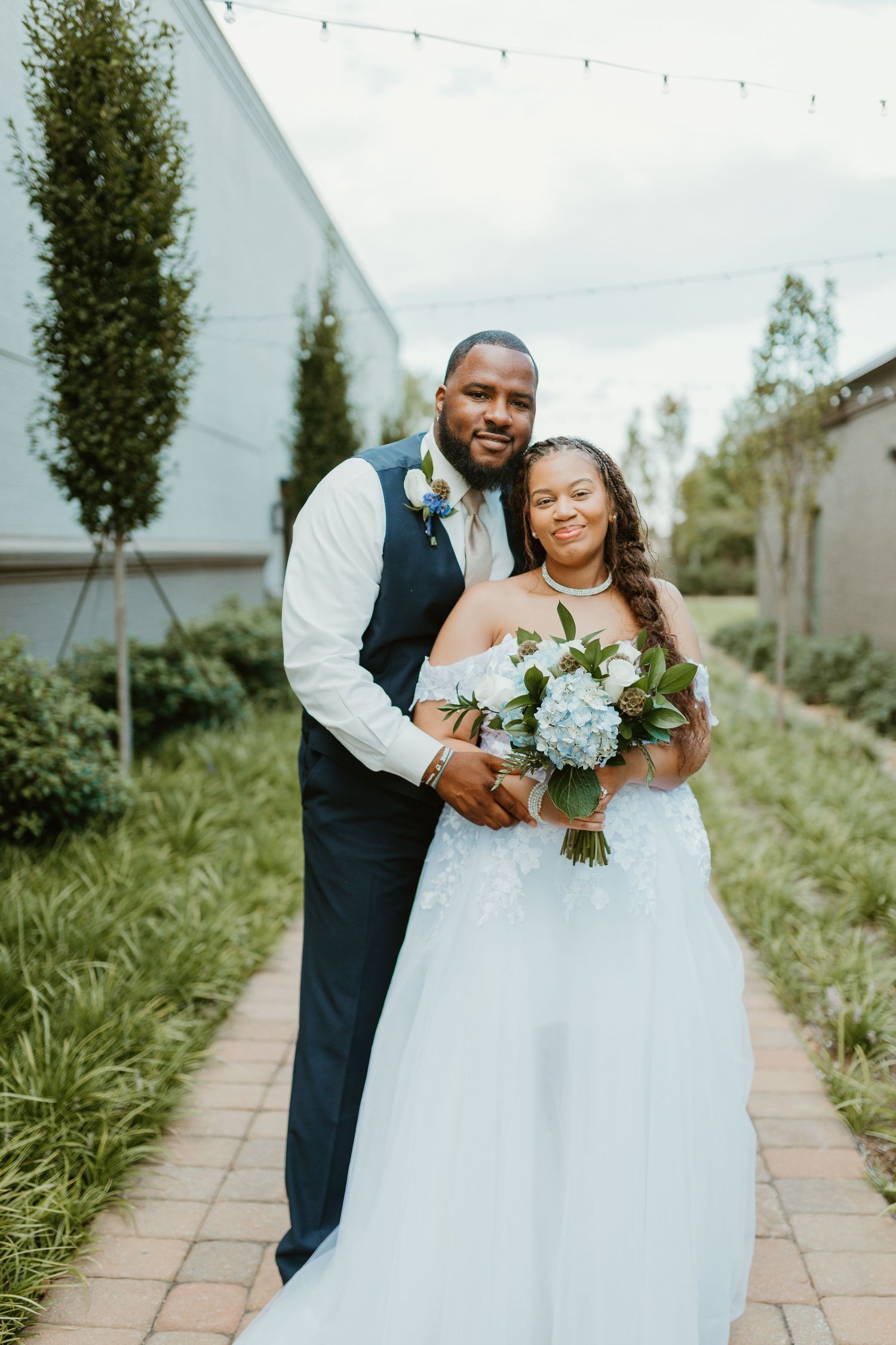 Newlywed couple embraces outdoors on stone path with blue floral bouquet and white wedding dress. Spartanburg SC