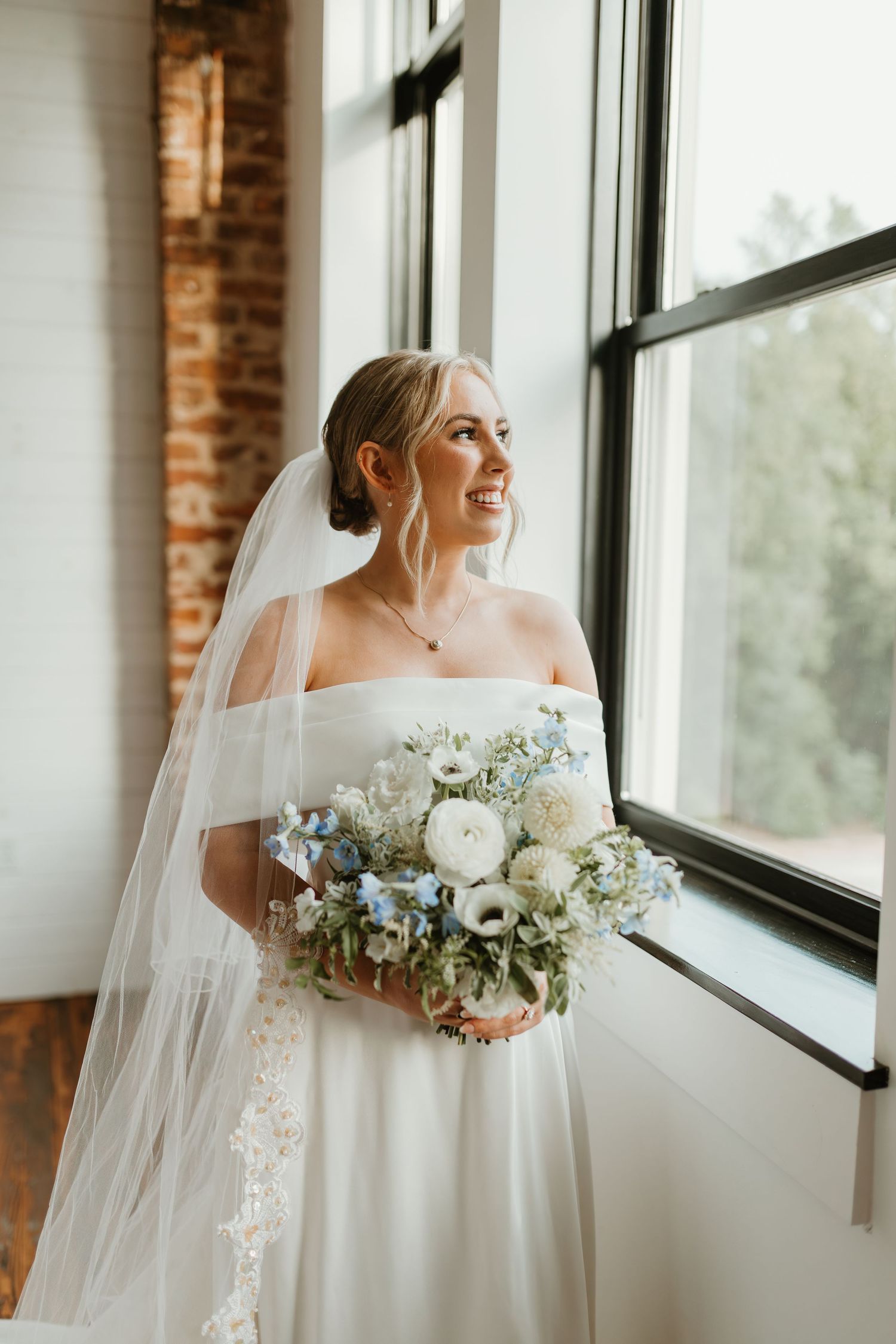 A bride in an off-shoulder white gown holds a blue and white bouquet while looking out a sunny window.