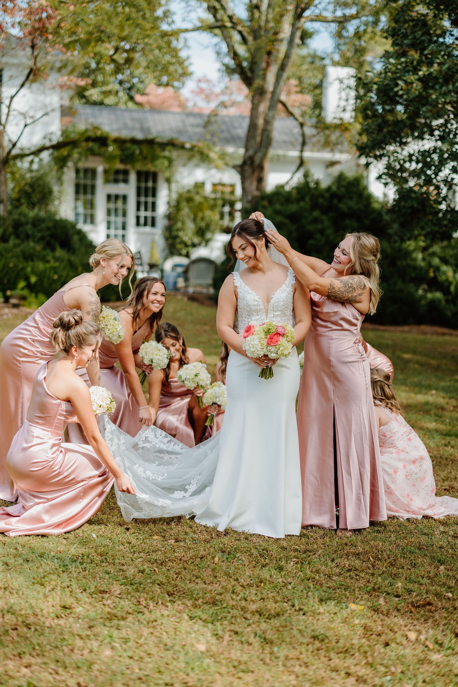 Wedding party in pink bridesmaid dresses poses playfully on grass during outdoor photo session.