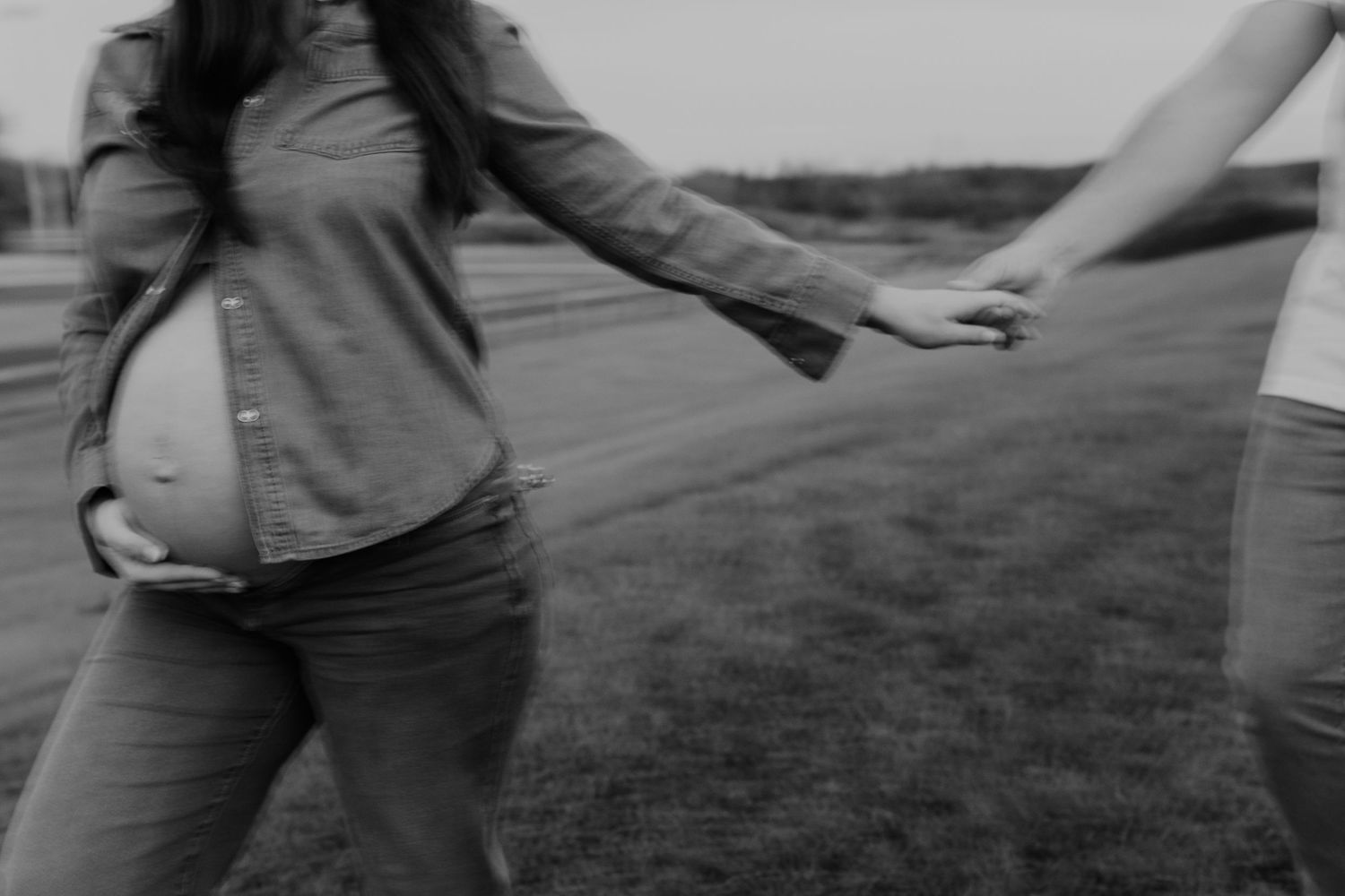 Black and white image of two people holding hands while walking in a grassy field, the girl in the photo is pregnant. Photosbyallii