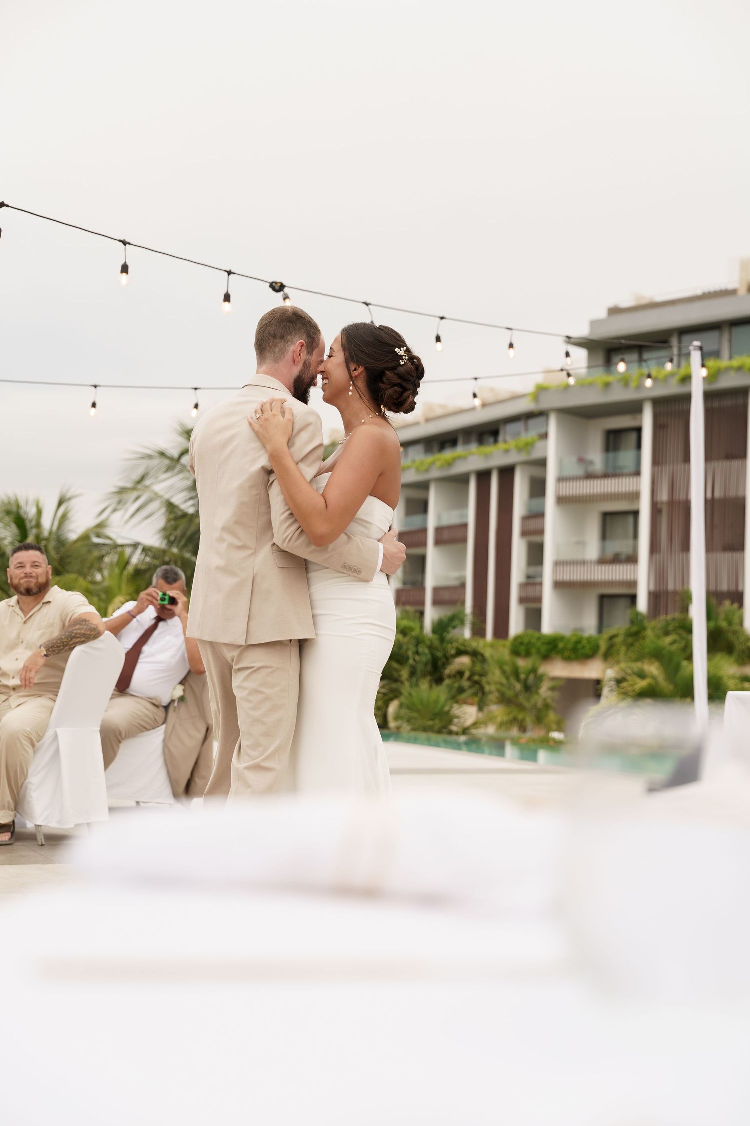 Romantic couples dance at an elegant outdoor wedding reception under string lights and palm trees.