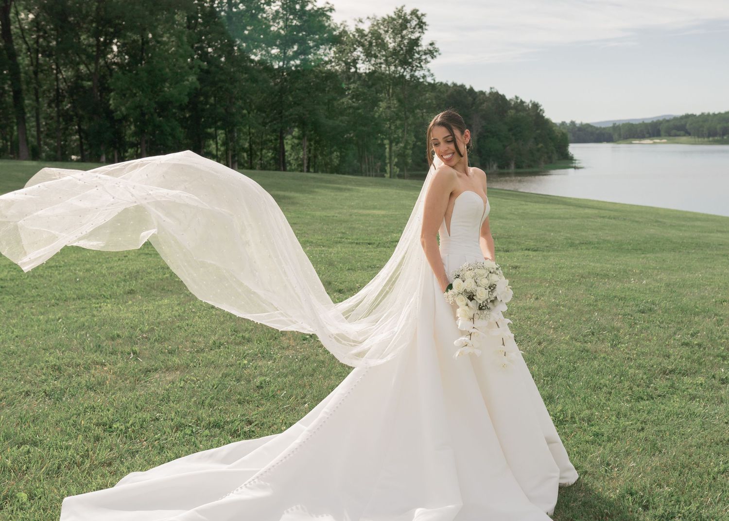 Bride in flowing white dress with long veil against lakeside landscape.