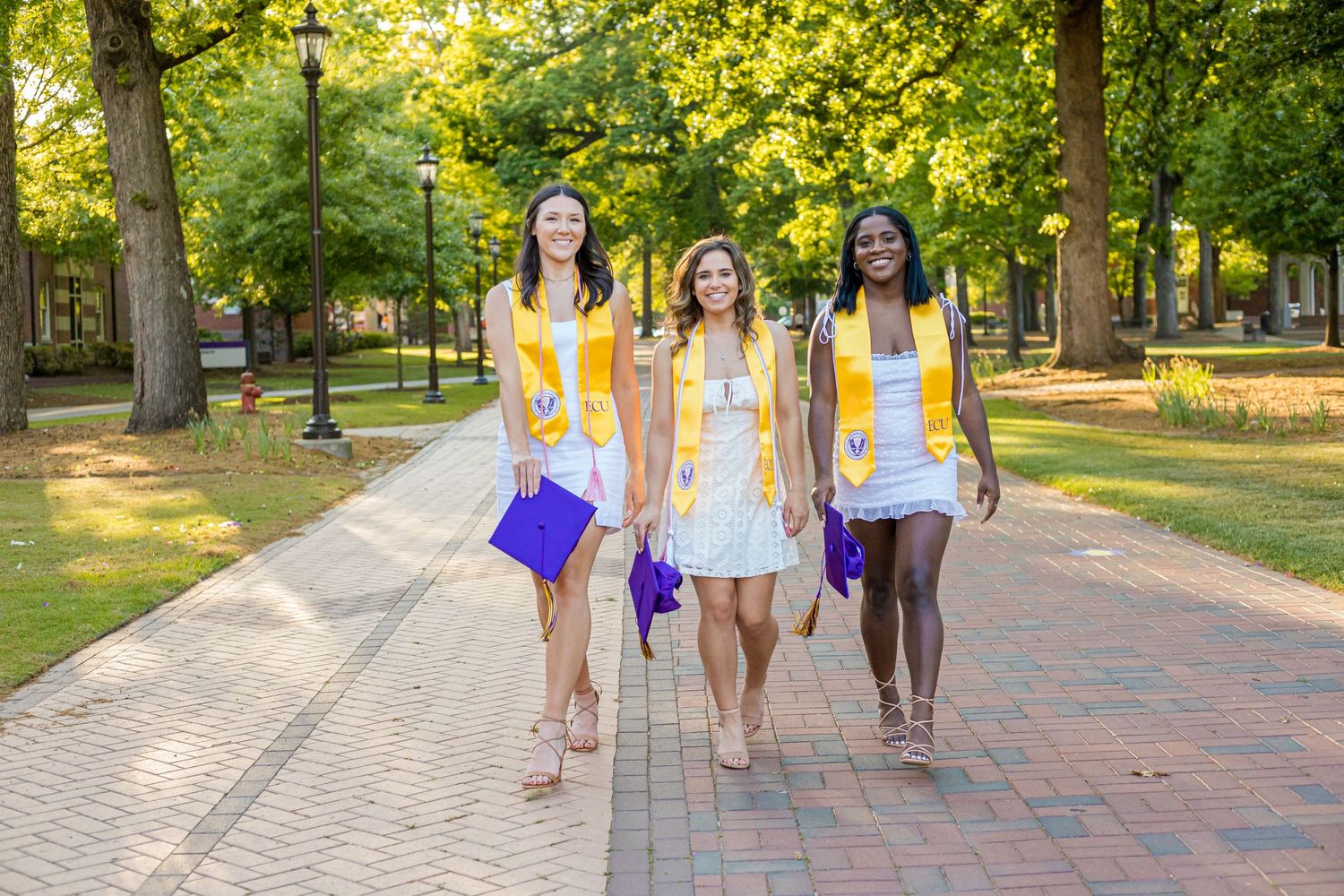 Three graduates in yellow stoles walk along a tree-lined brick path on their campus.