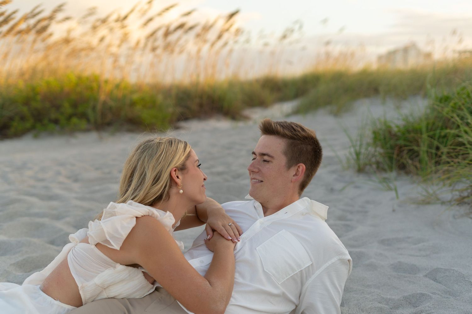 A couple in wedding attire embraces on a sandy beach at sunset during their romantic photoshoot.