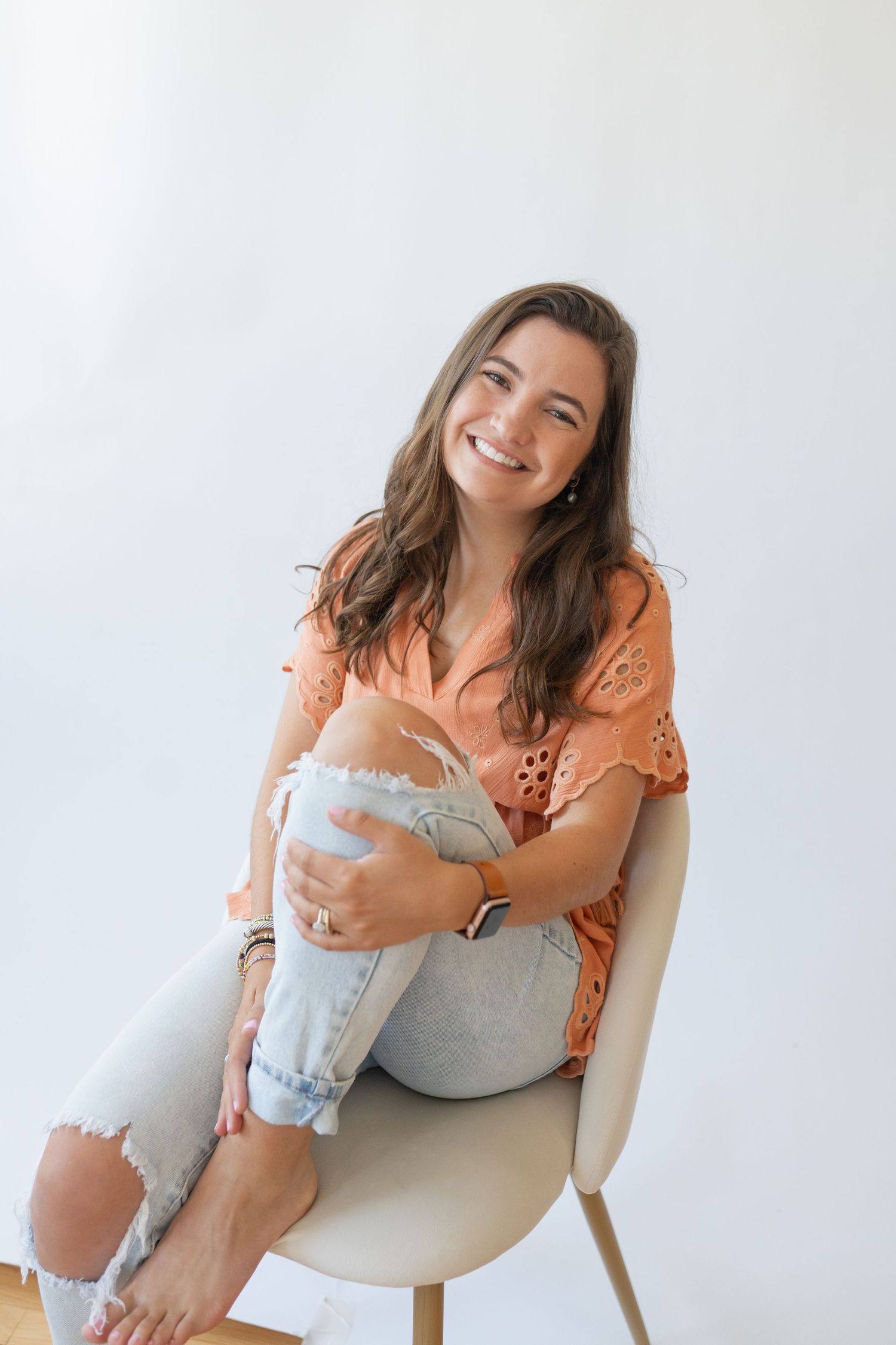 Woman in orange eyelet top sits casually on modern chair wearing ripped jeans.
