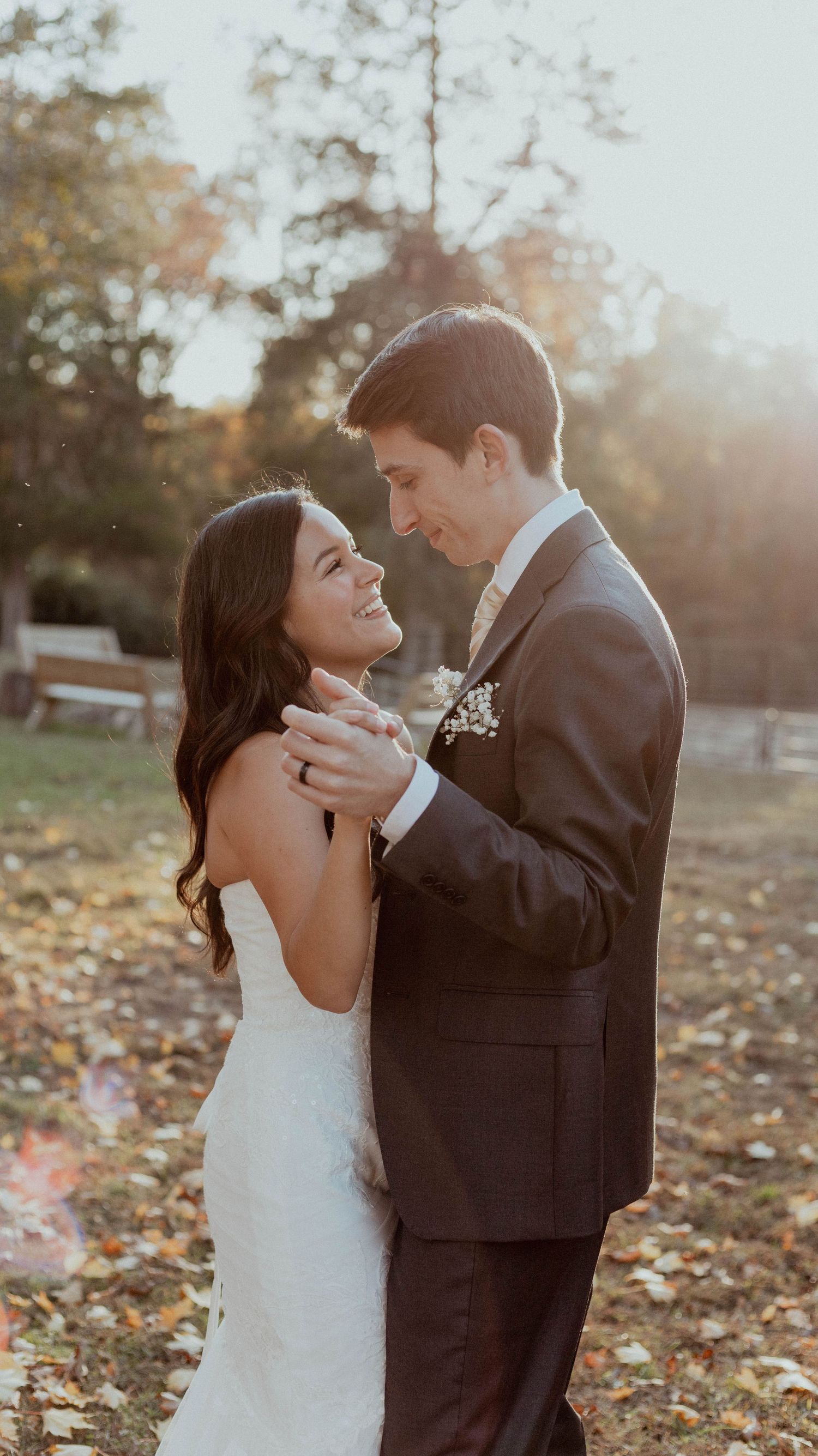 A romantic wedding couple embraces outdoors during golden hour with autumn leaves scattered on the ground.