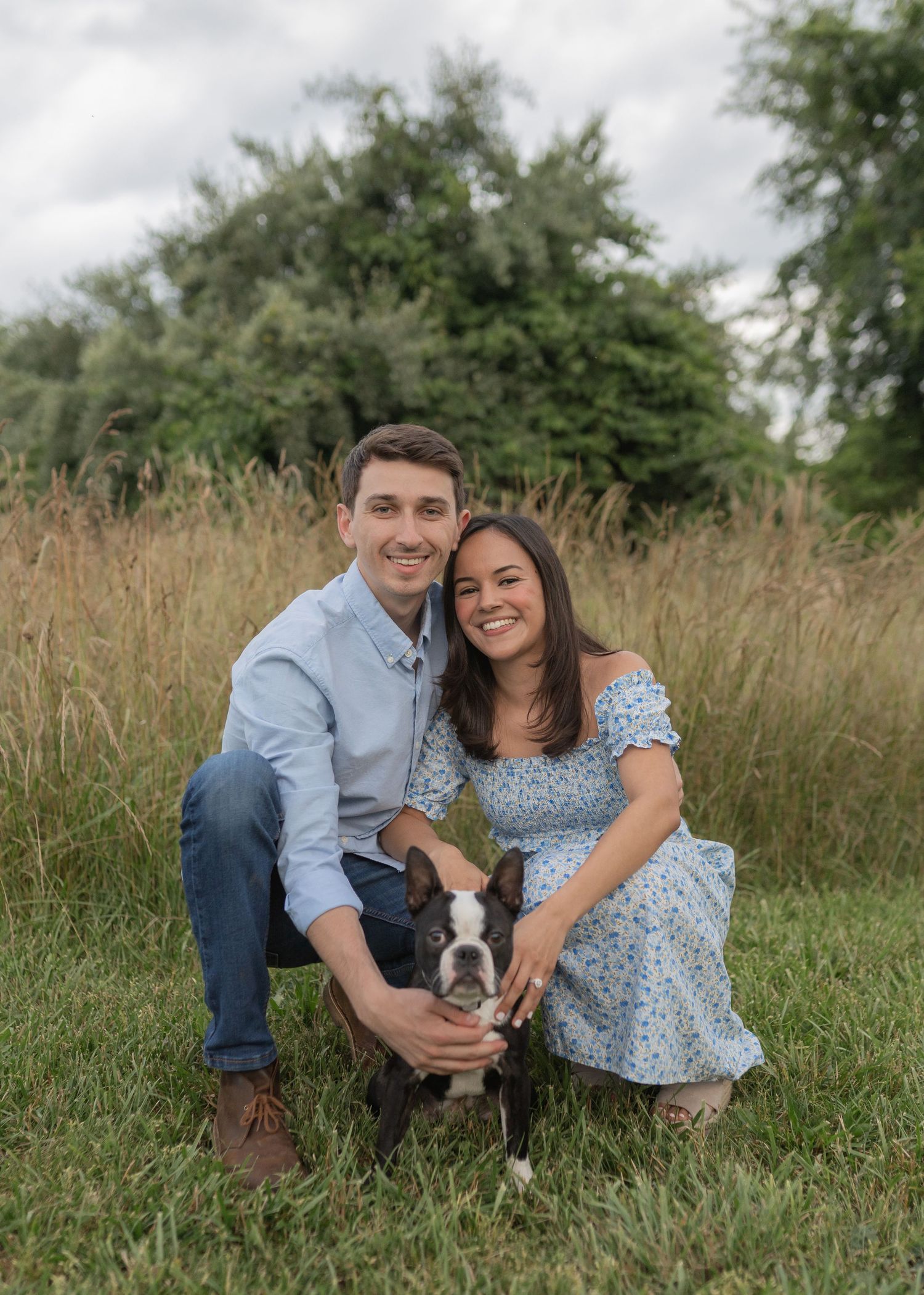 Couple sits in tall grass field with their small dog while wearing coordinated blue and white outfits.