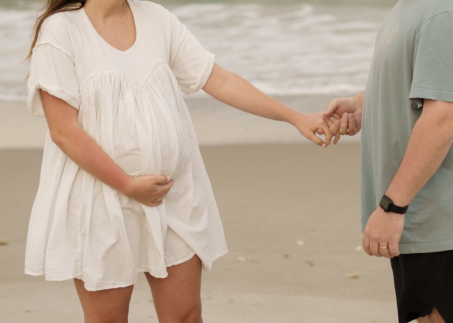 Couple holding hands on a beach, with a flowy white dress visible against the sand.