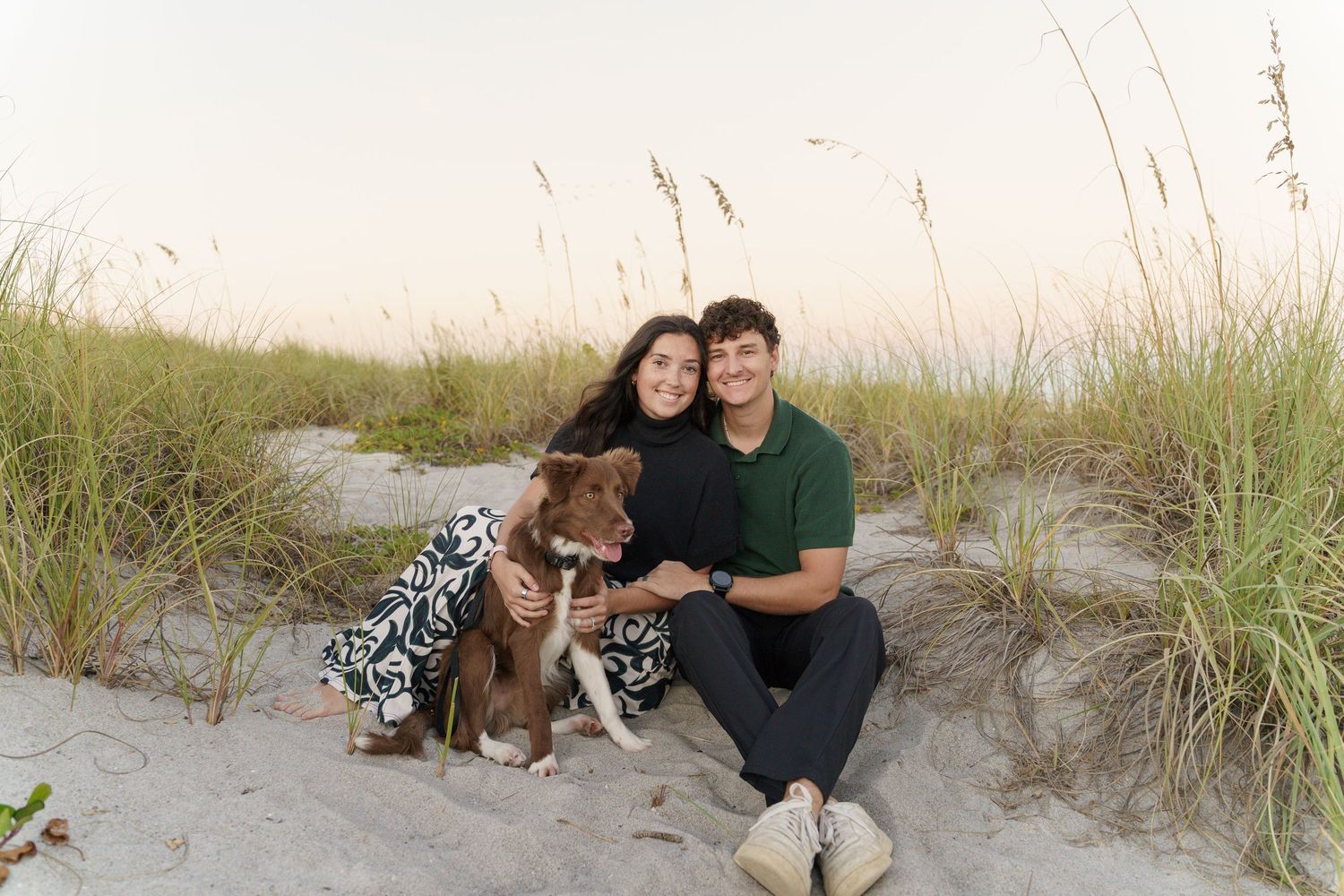 A group of people and a dog pose together on a sandy beach with tall grass during sunset.